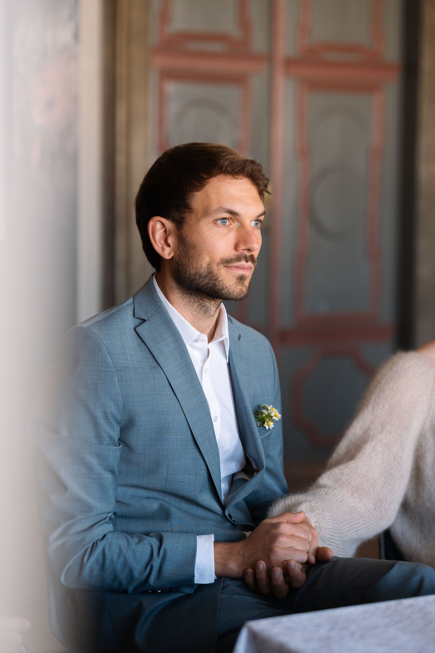 A well-dressed man in a blue suit with a floral pocket square sits thoughtfully in an elegant interior setting.