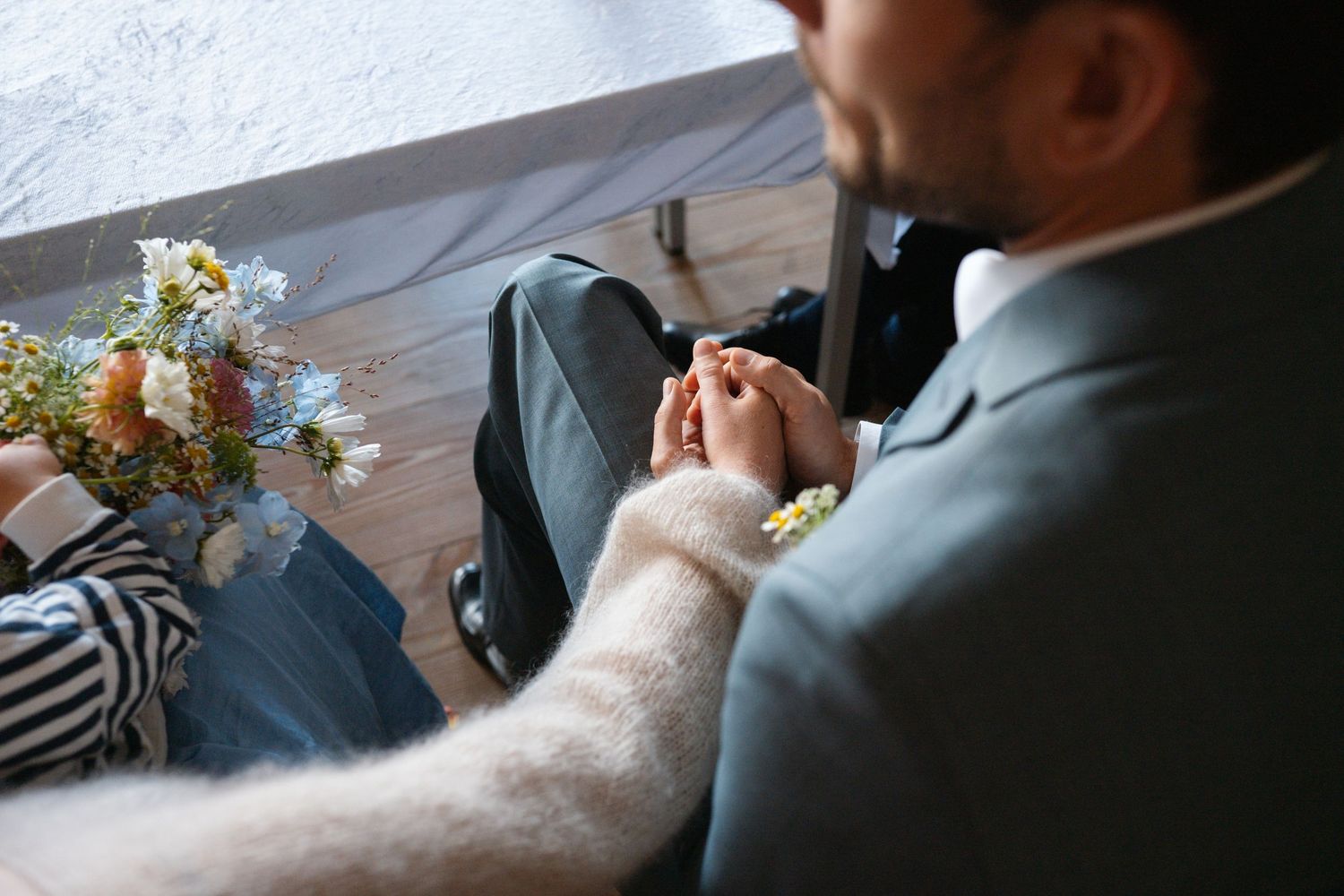Couple holding hands tenderly at a wedding ceremony, with a bouquet of wildflowers nearby.