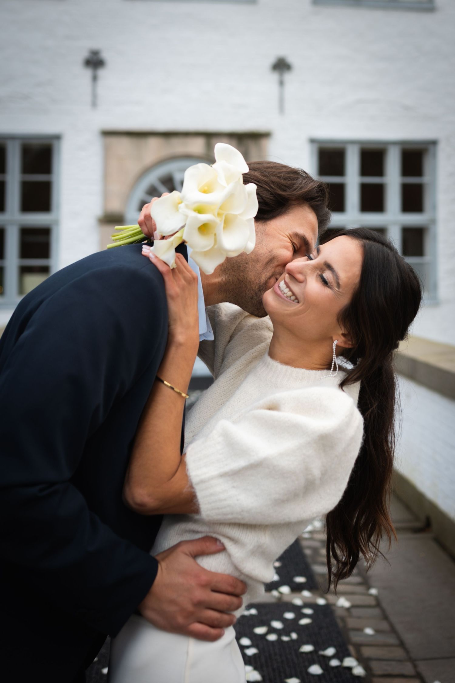 A couple shares a playful moment leaning back while holding white flowers between their smiles.