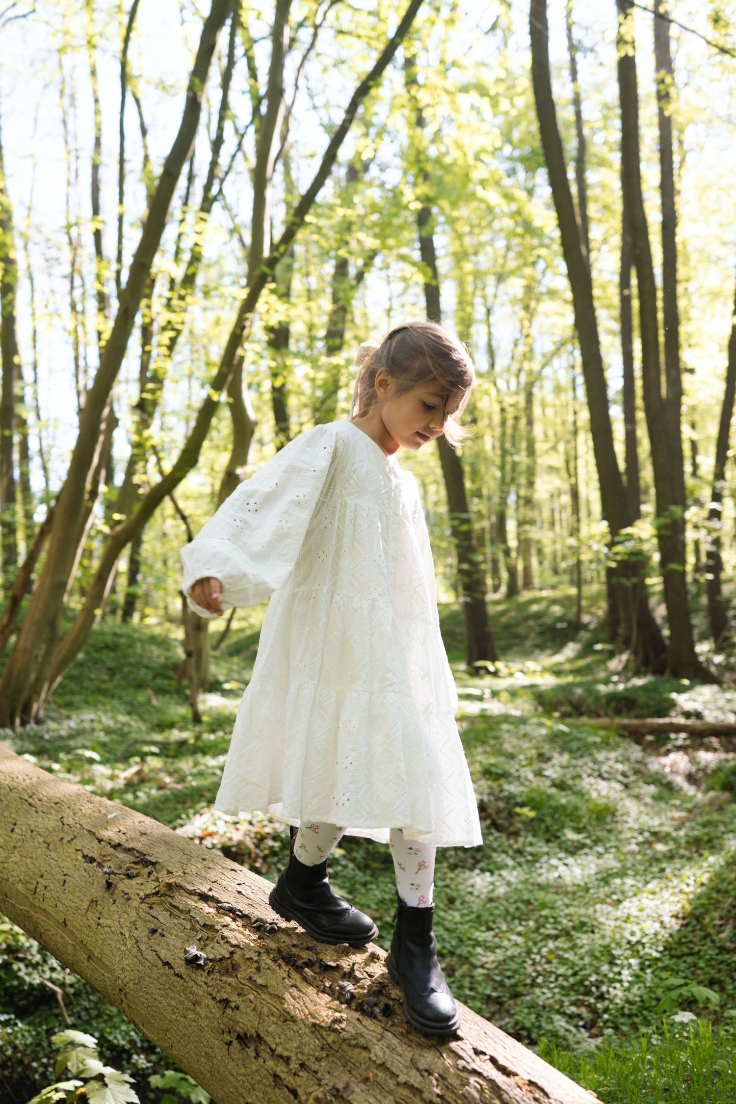 Young girl in white dress and black boots balancing on a fallen log in a sunlit forest.