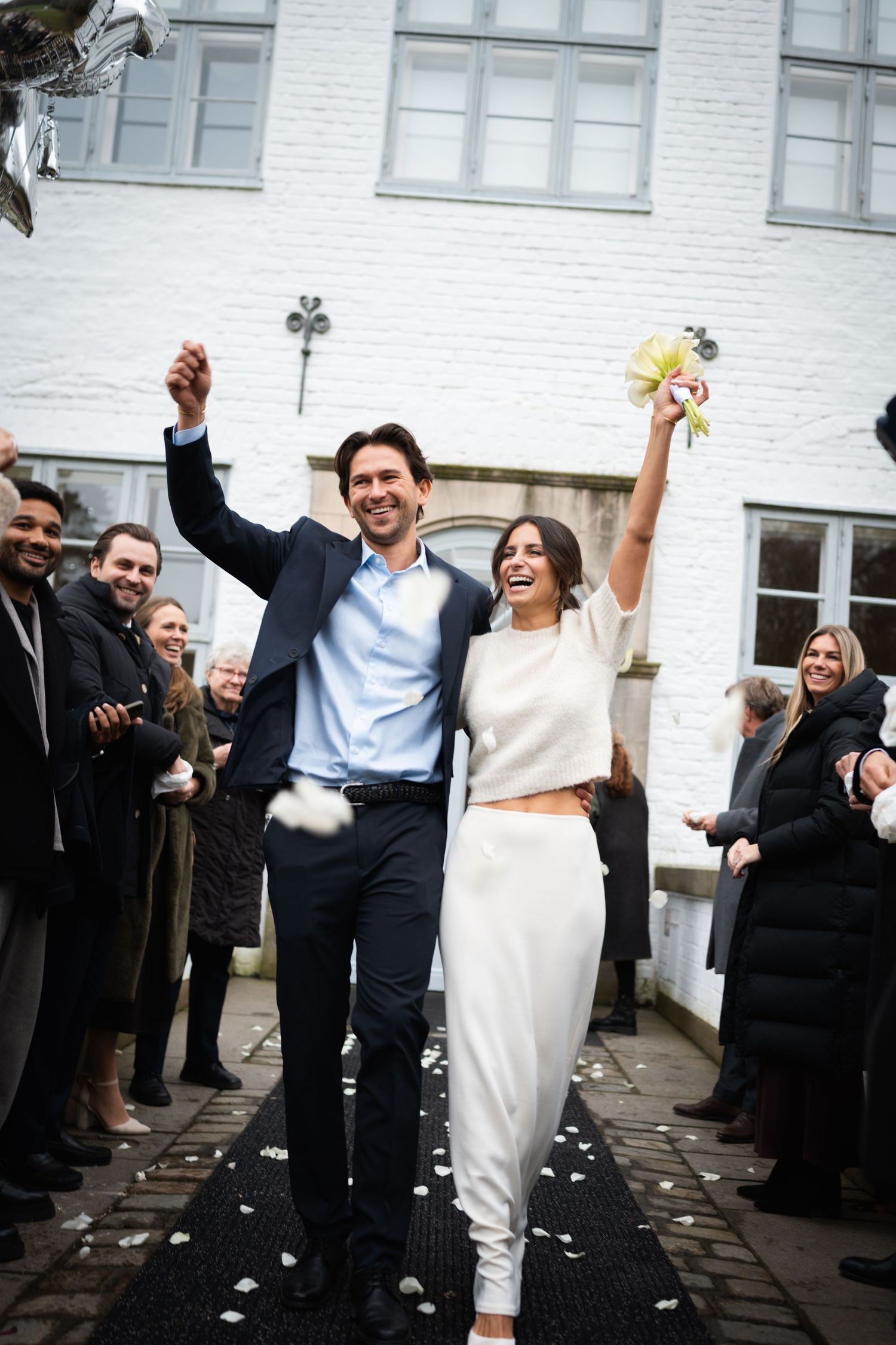 Joyful newlywed couple celebrating after wedding ceremony, walking through cheering guests outside white brick building.