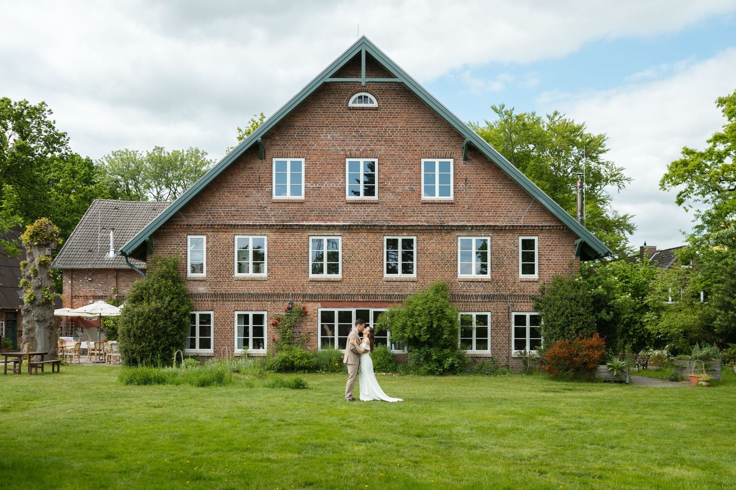 Bride and groom standing on lush green lawn in front of a large brick wedding venue surrounded by trees.