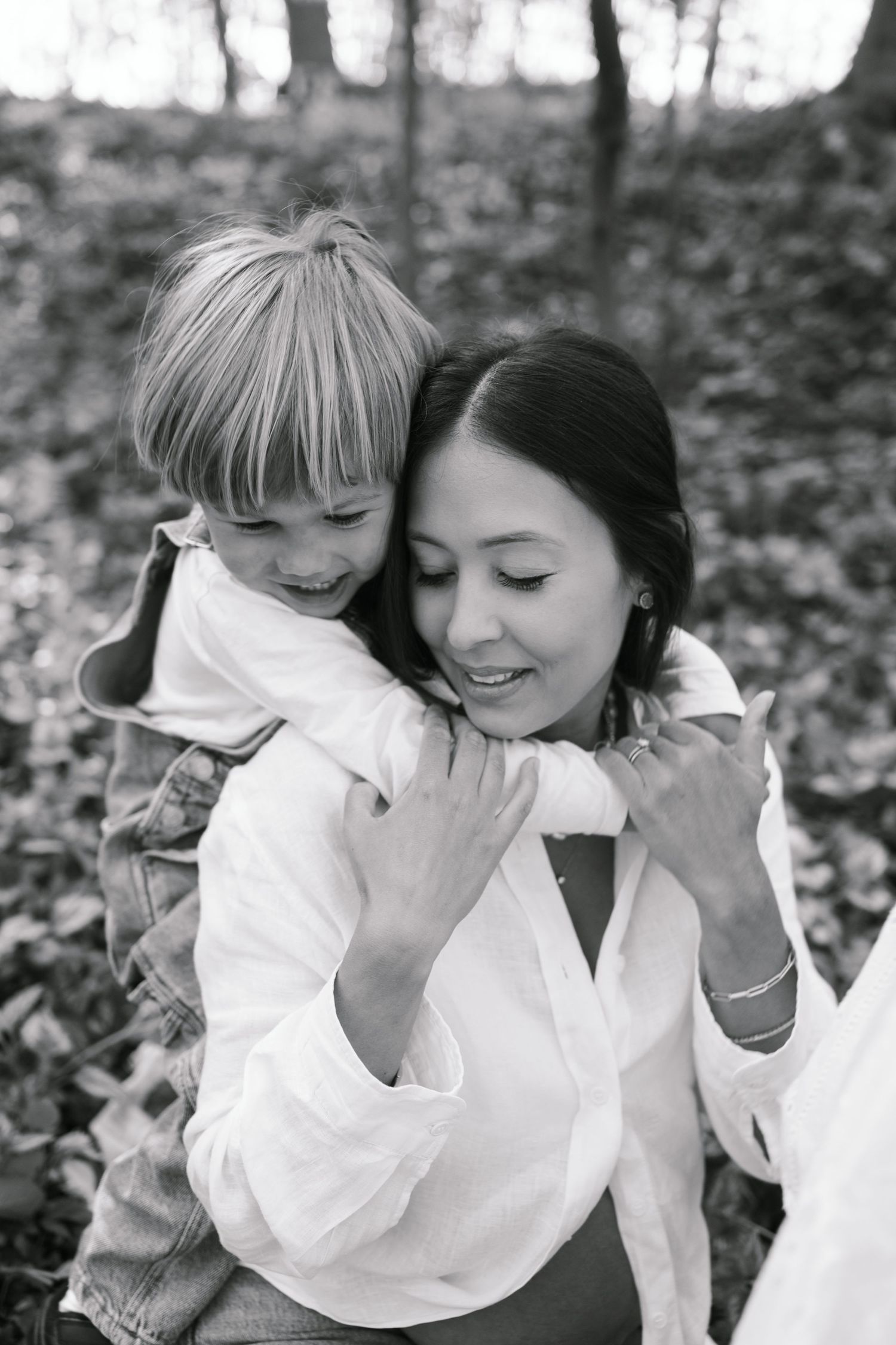 Black and white photo of a smiling mother hugging her young blonde child outdoors among fallen leaves.