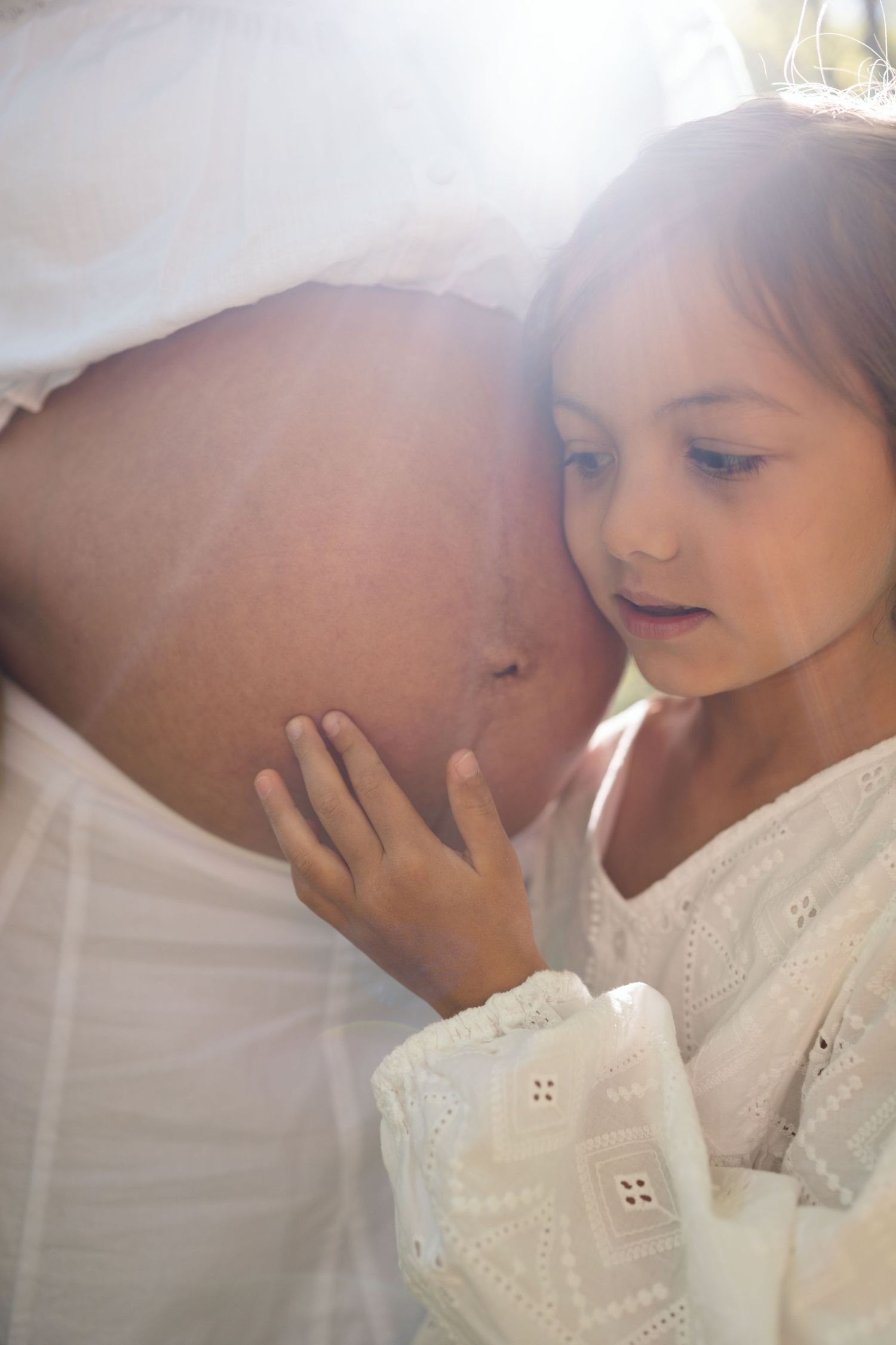 Young girl in white dress listening to pregnant mother's belly in soft natural sunlight.