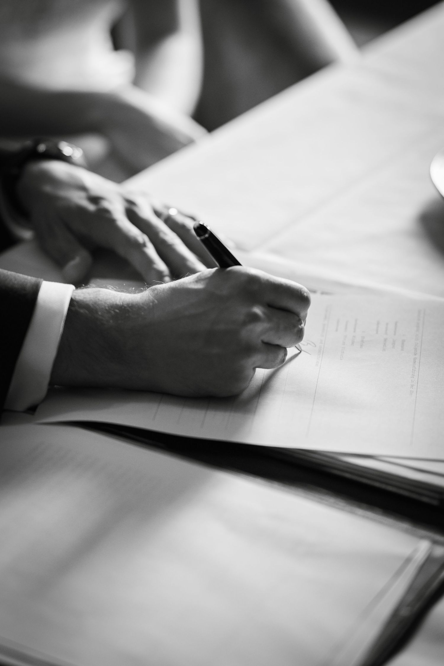 Close-up black and white image of hands writing or studying with papers and documents on a desk surface.