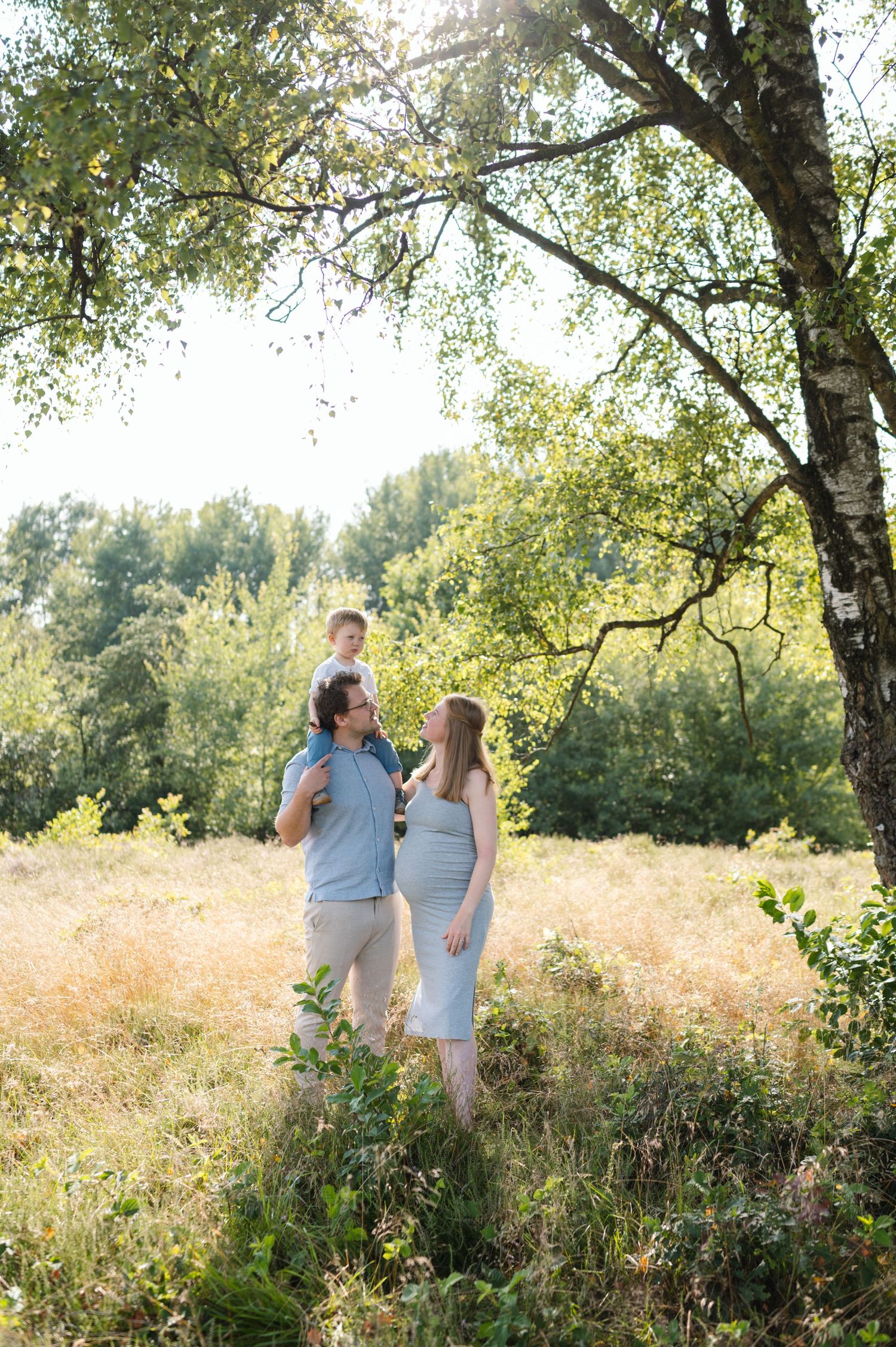 A couple shares tender moments in a sunlit meadow surrounded by trees during a romantic outdoor photo session.