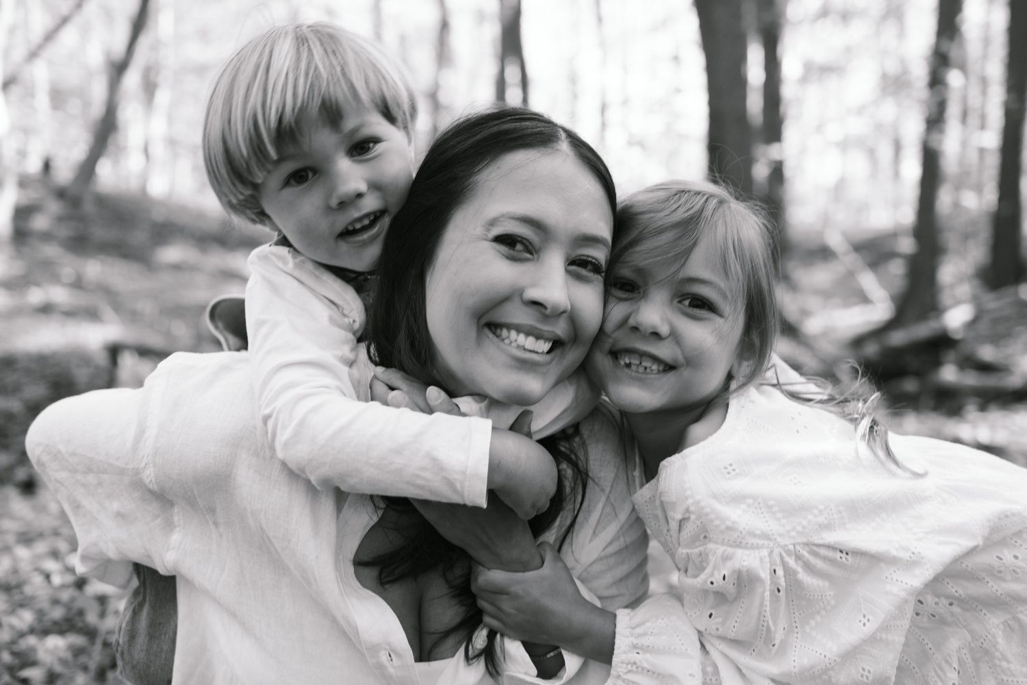 Black and white photo of a smiling mother hugging two young children in a forest setting.