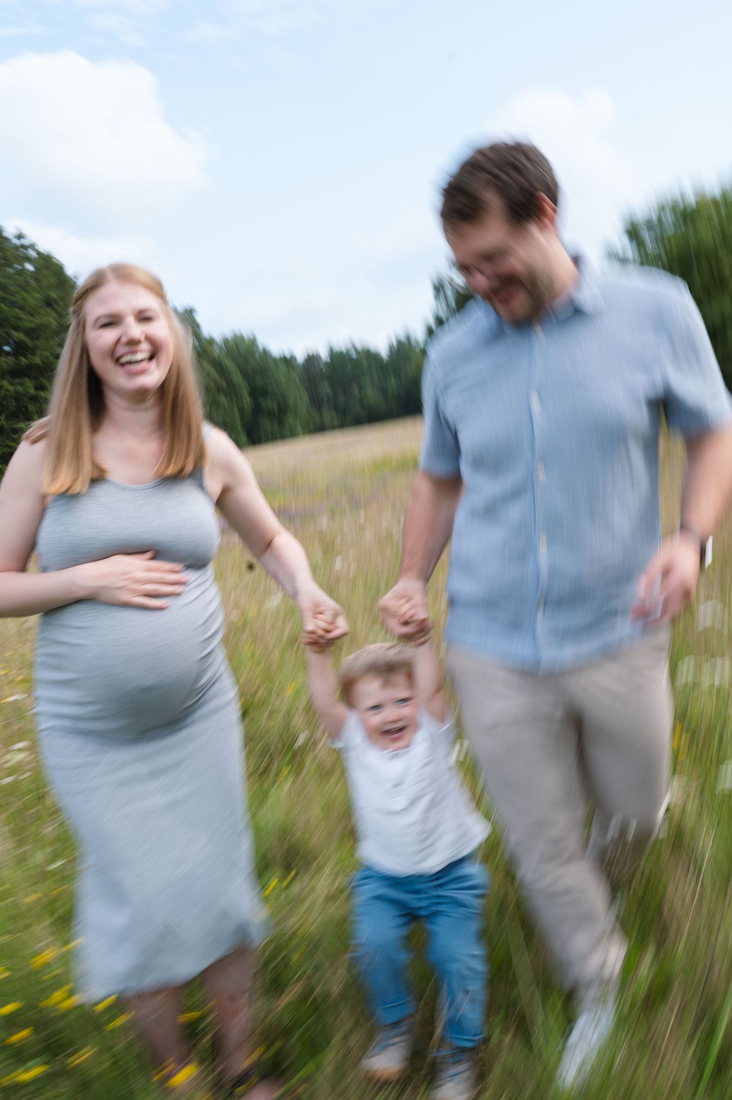 A family runs and plays together in a sunny meadow filled with tall grass and wildflowers.