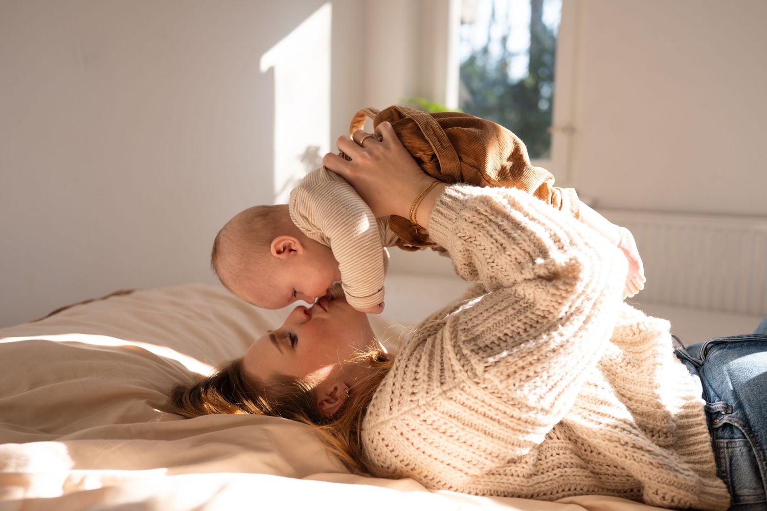 Mother lying on bed lifting her baby in the air while sharing a tender kiss in warm sunlight.