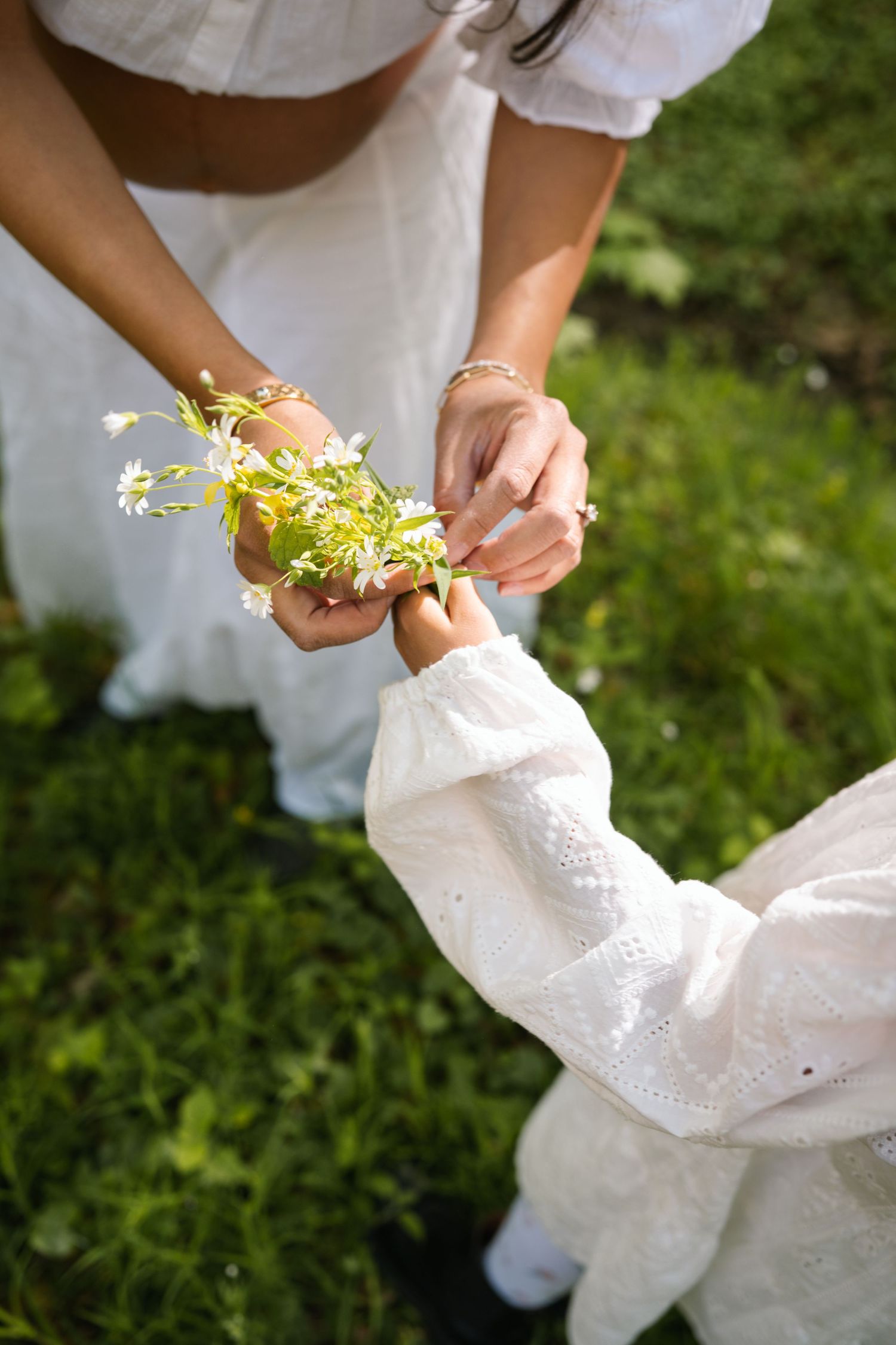 Adult and child in white dresses sharing a small bouquet of wildflowers on green grass.