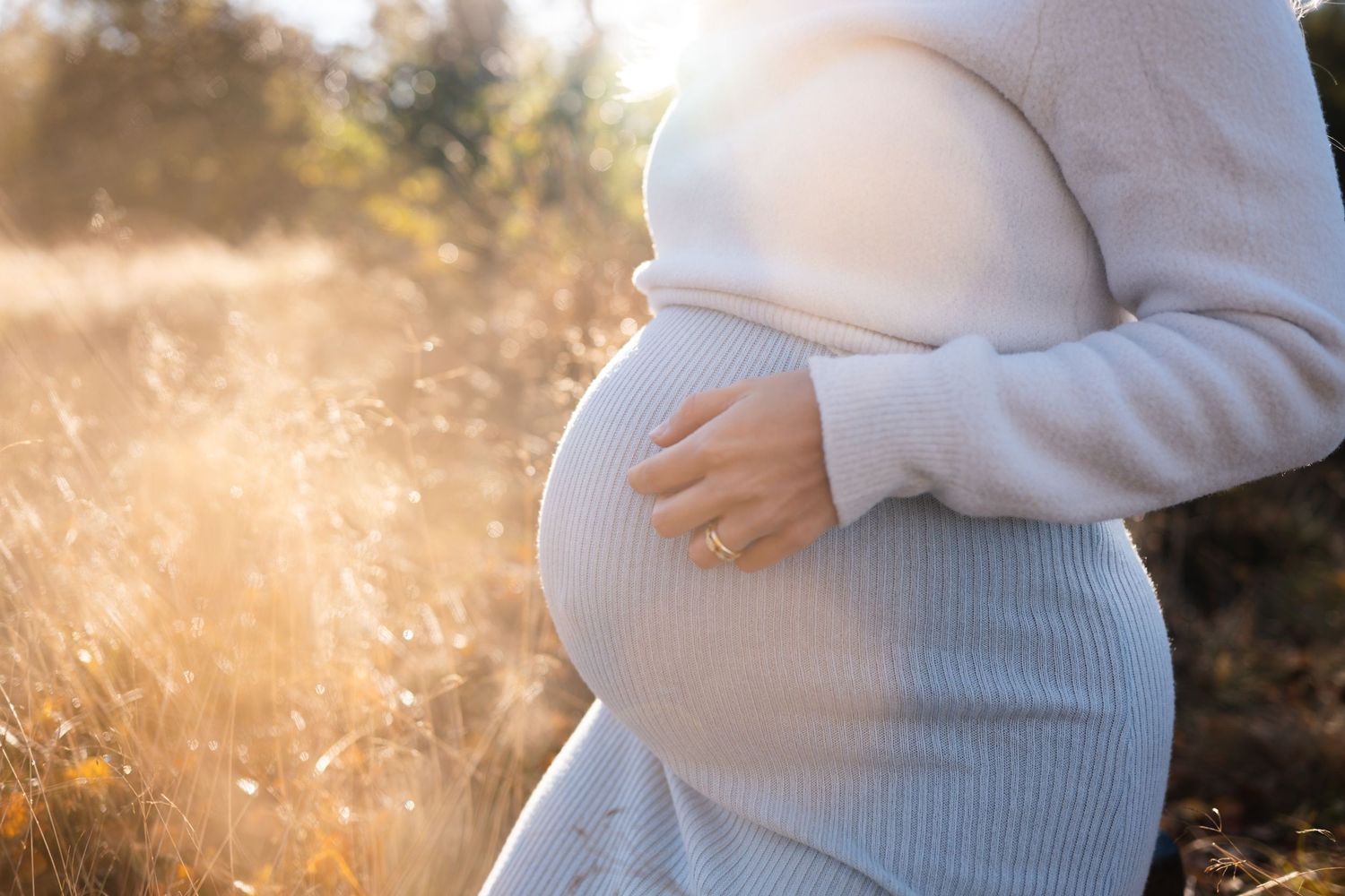Pregnant woman in white sweater cradles belly during golden hour sunset with sunlit grass in background.