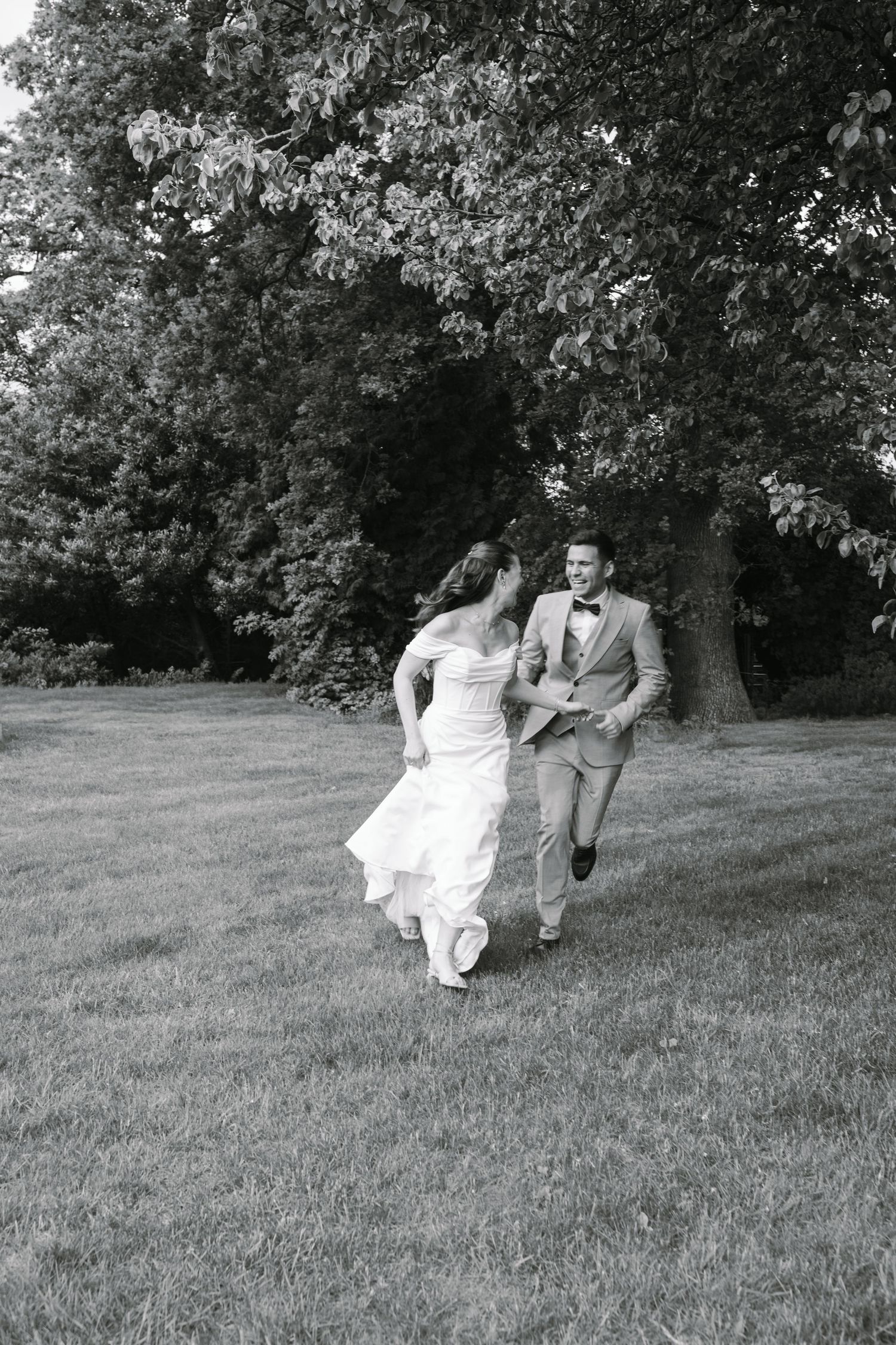 Black and white photo of a bride and groom running and laughing together across a lush green lawn.