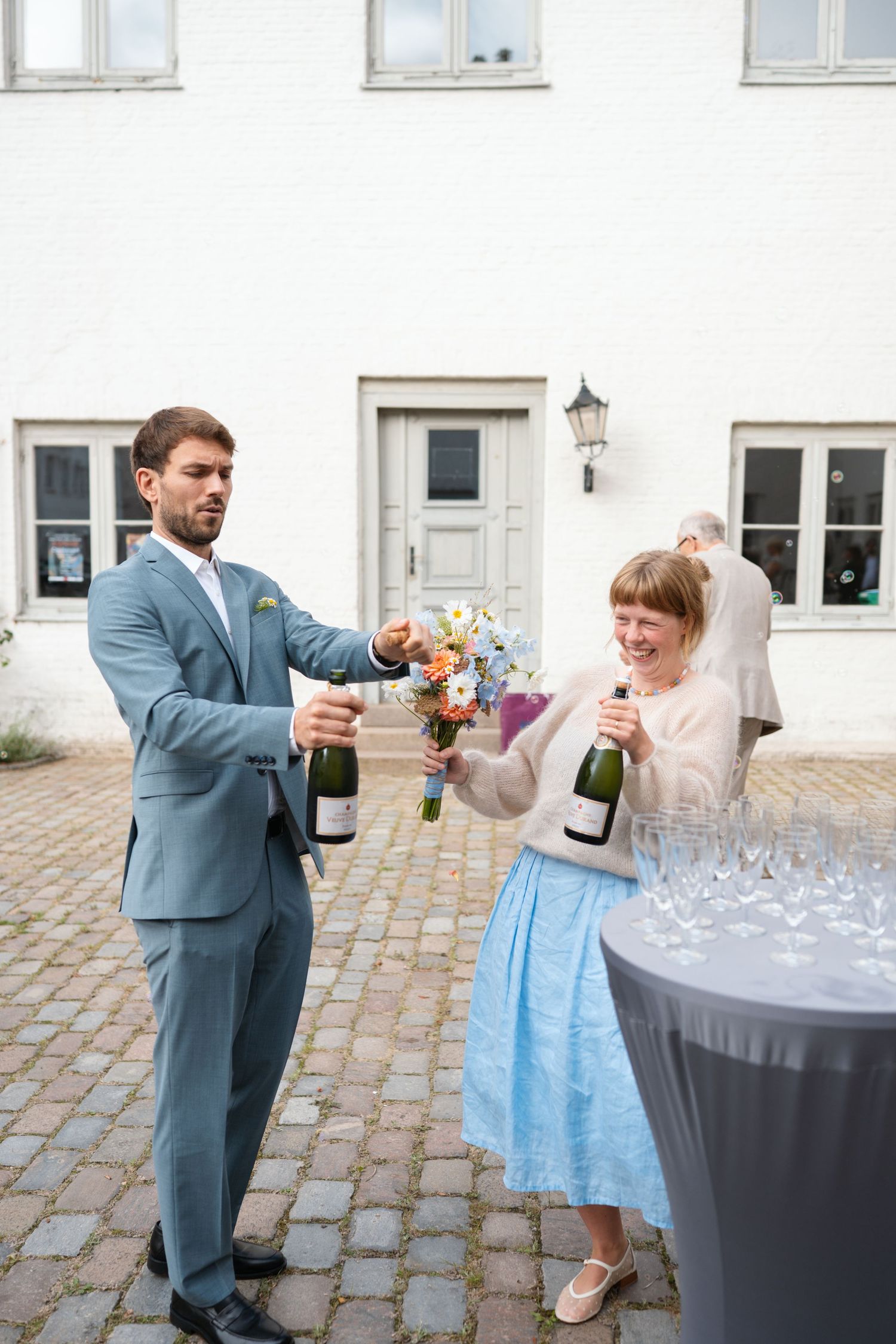 Bride and groom opening champagne bottles outdoors at wedding venue, bride holding colorful bouquet wearing blue dress and veil.