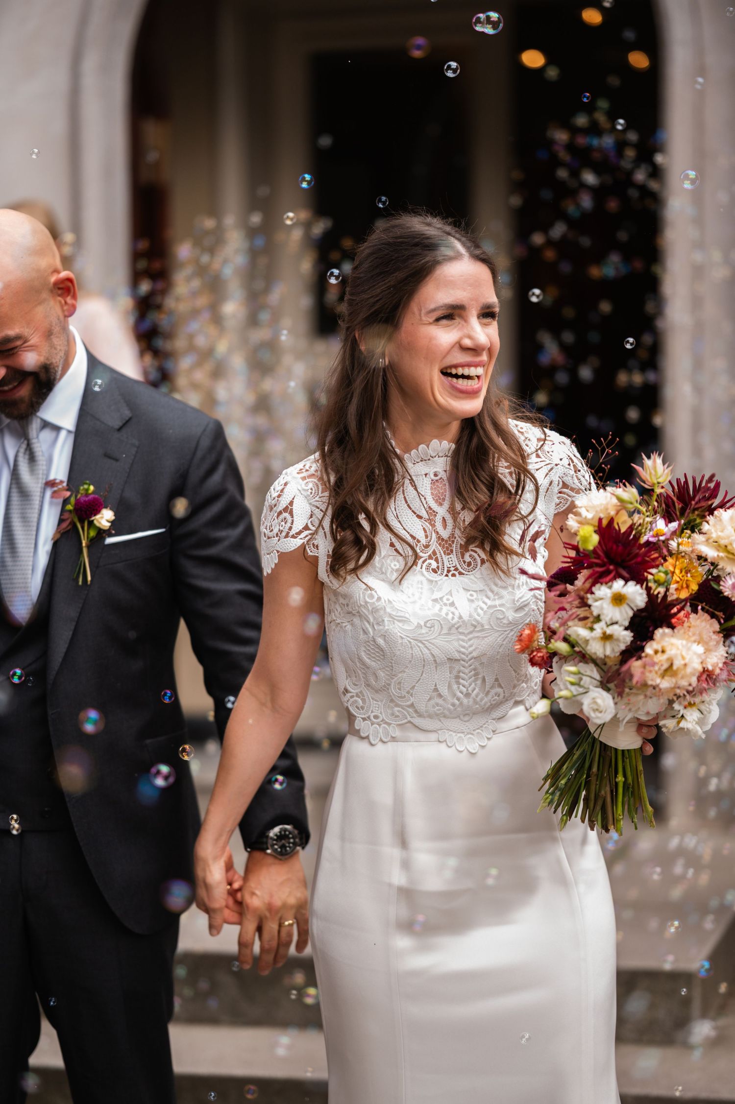 Happy bride laughing while holding autumn bouquet with burgundy dahlias and blush roses during confetti toss celebration.