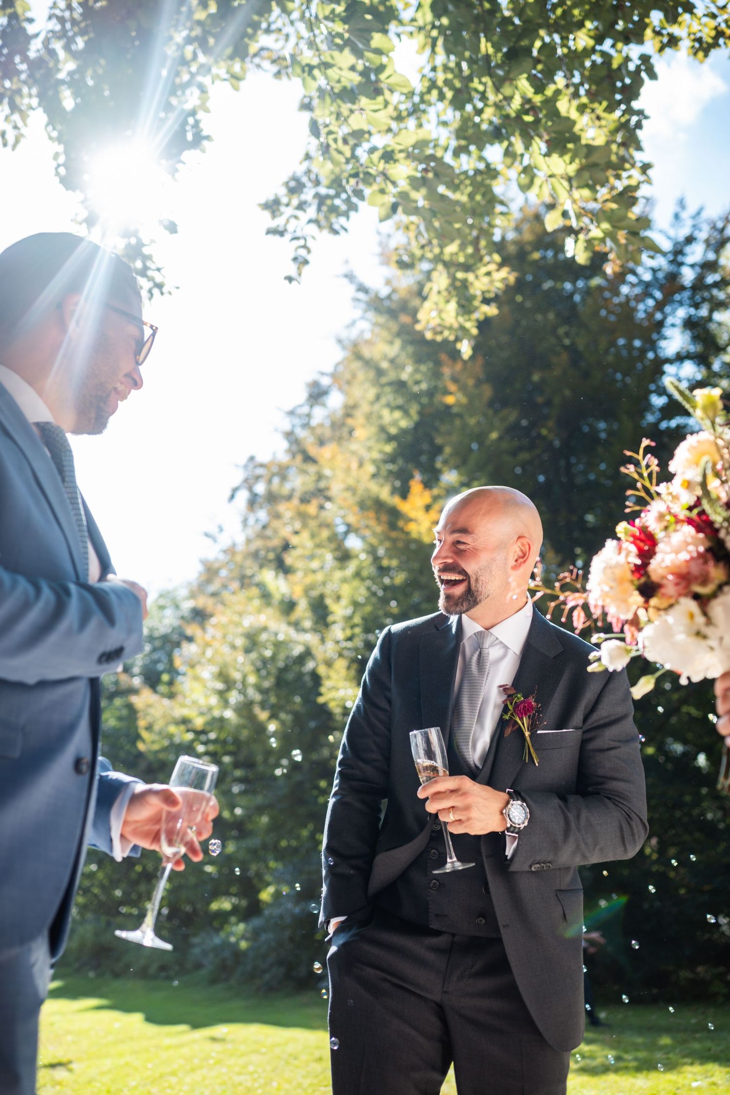 Wedding guests in formal attire share laughter and champagne during an outdoor garden reception celebration.