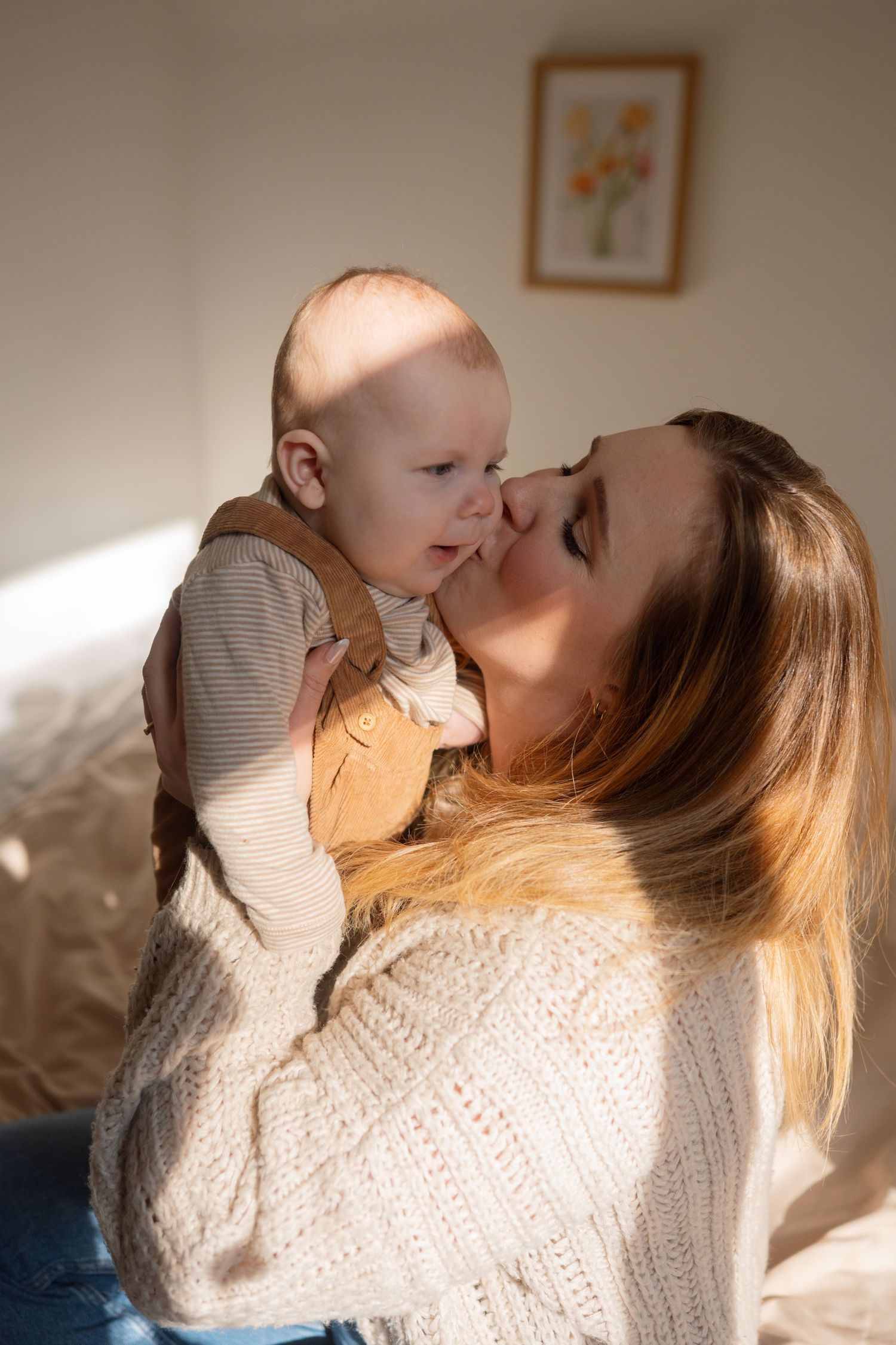 A loving mother holds her baby close for a gentle kiss in warm sunlit bedroom.