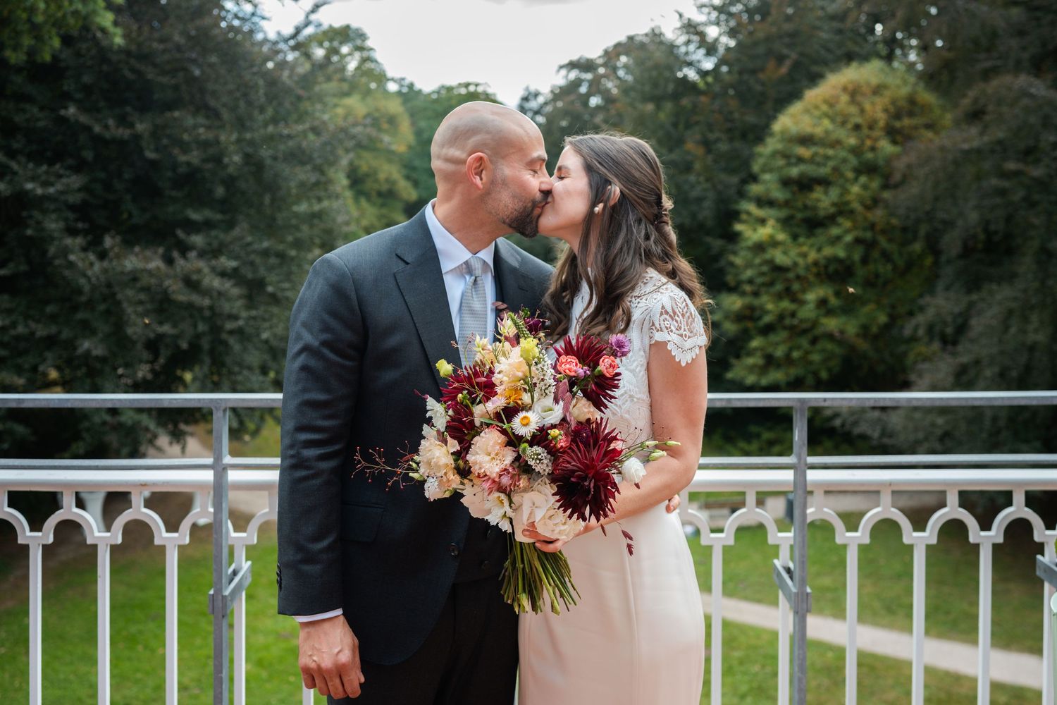 A romantic moment shared between a couple on a white railing bridge surrounded by lush greenery while holding a floral bouquet.
