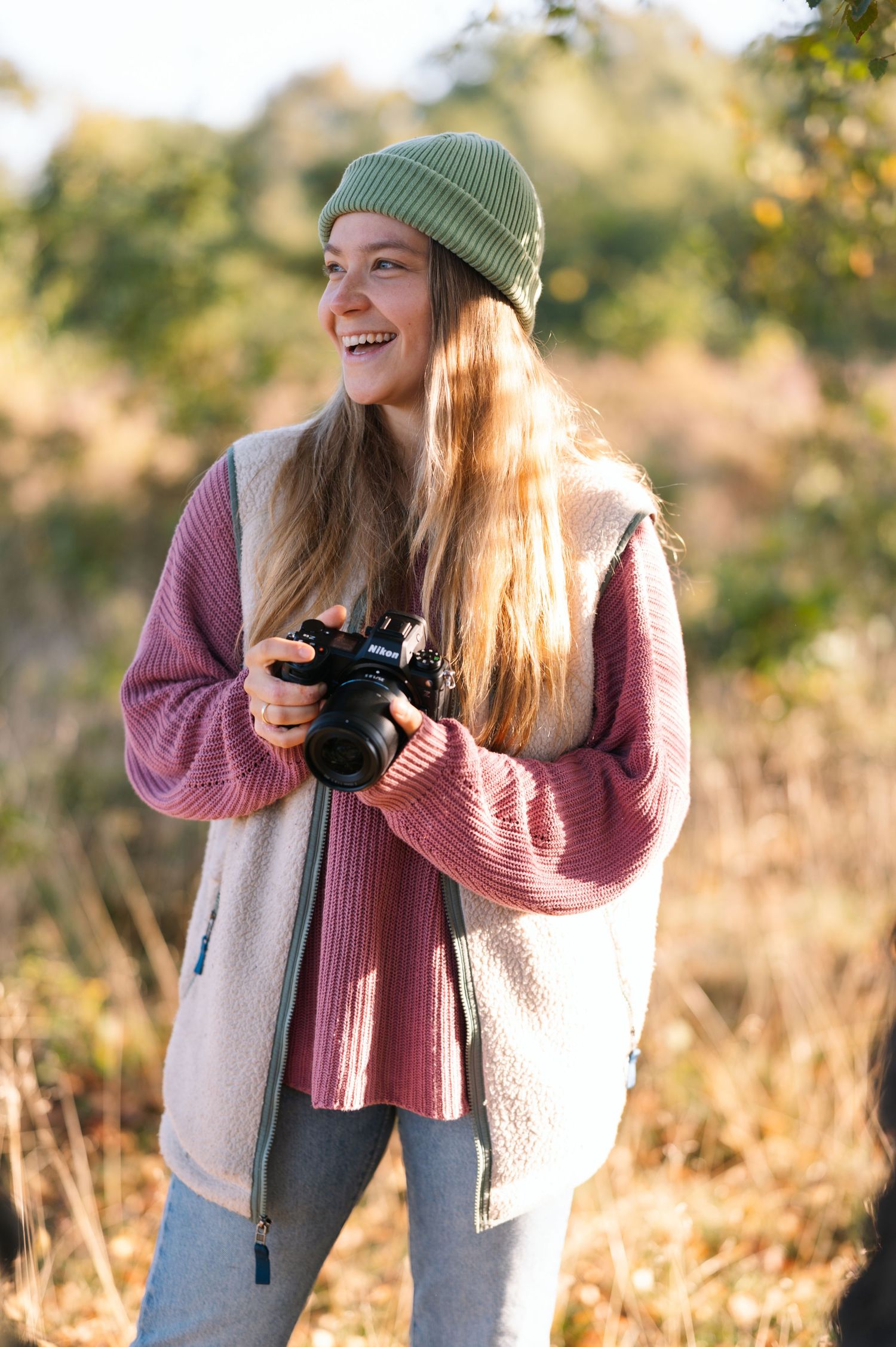 Smiling young woman holding a camera outdoors in autumn, wearing a green beanie and pink sweater.