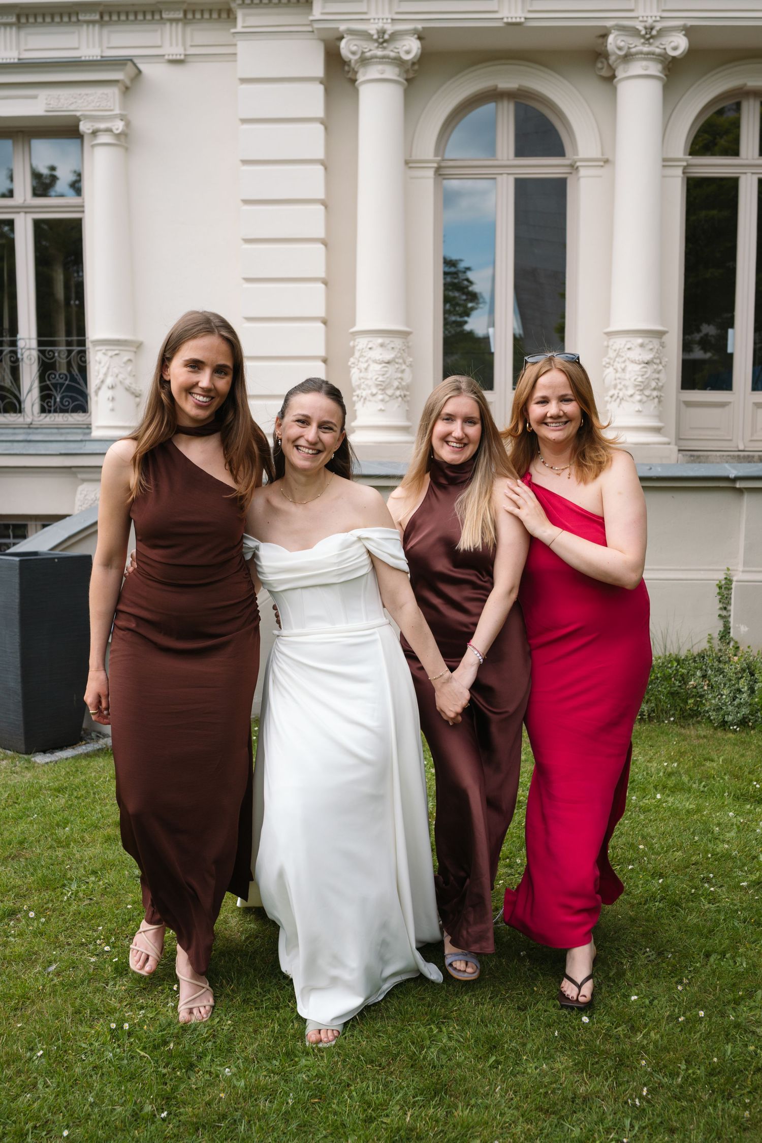 A smiling bride in a white gown poses with three bridesmaids in brown and red dresses outside a venue.