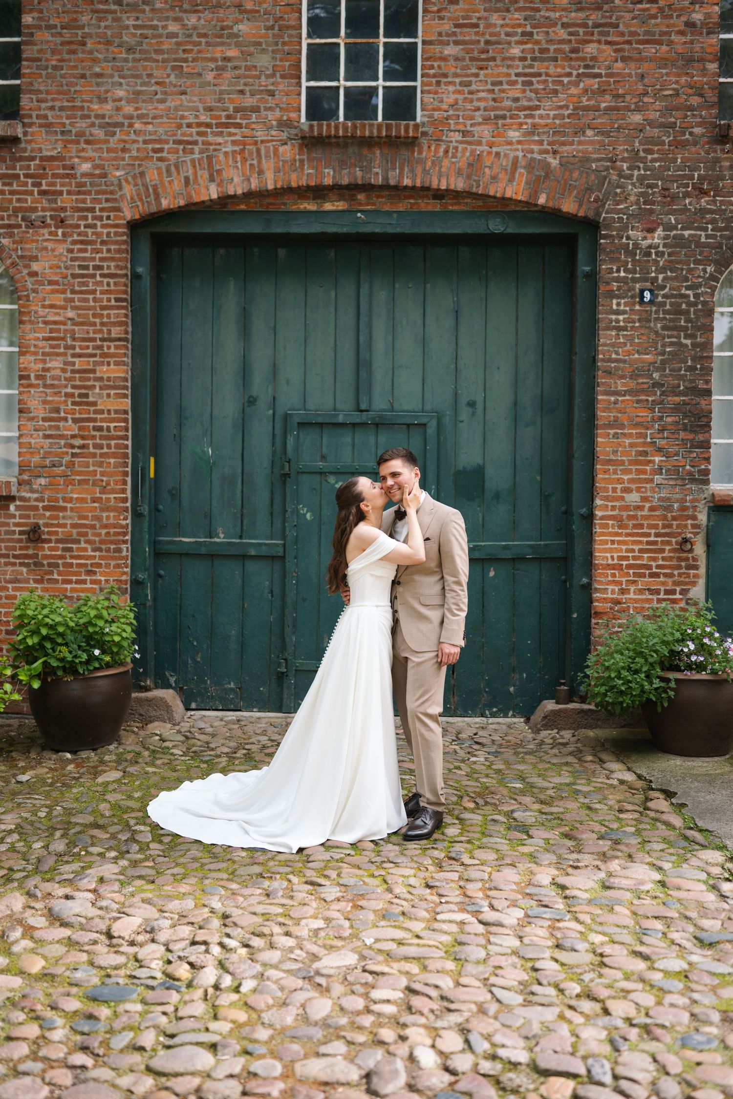 Bride and groom embrace in front of a rustic green wooden door on cobblestone, with brick wall backdrop.