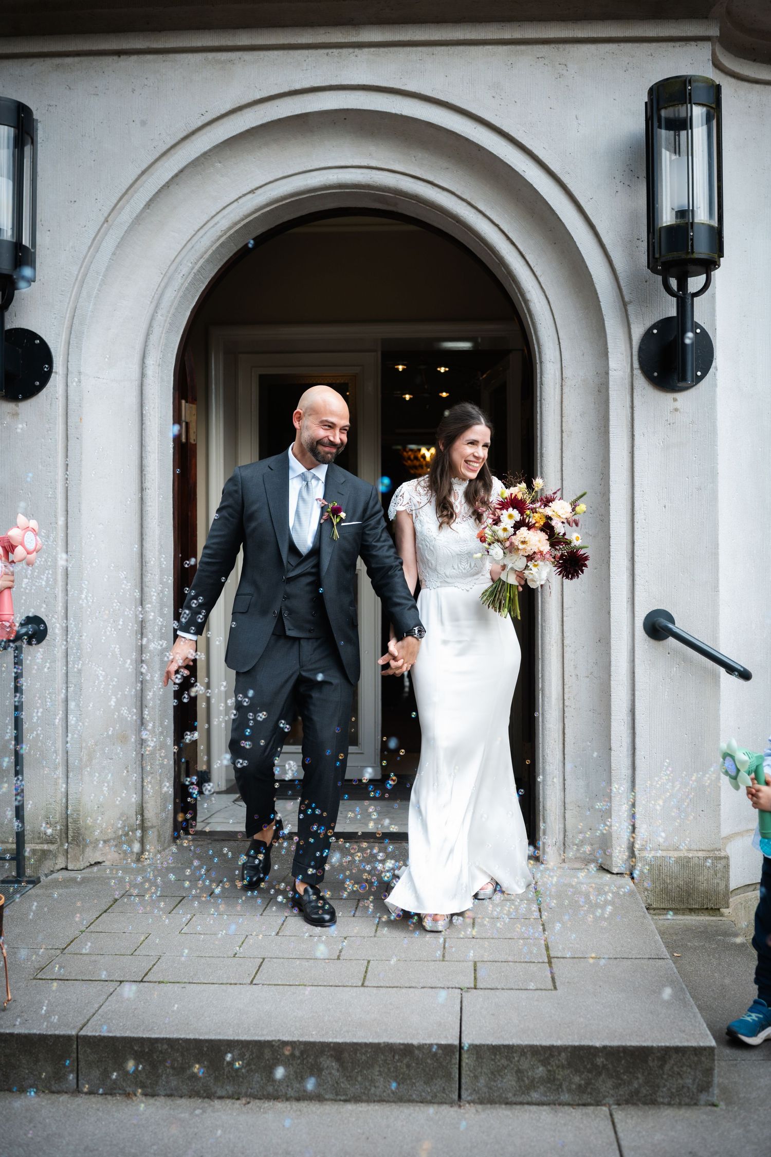 A wedding couple runs through an arched stone colonnade while guests throw confetti and flower petals in celebration.