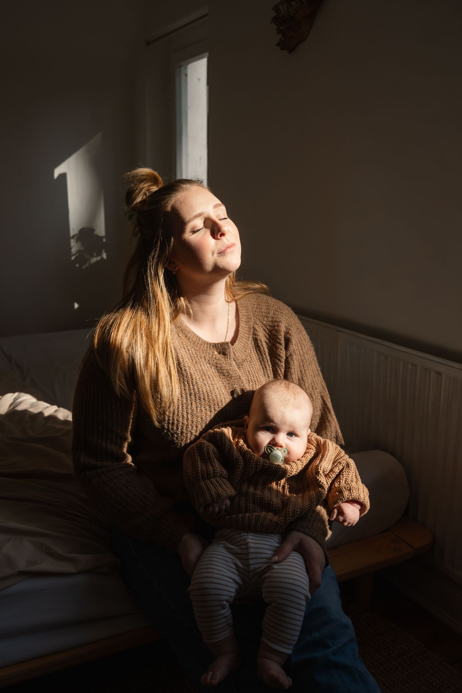Mother holding baby in warm sunlight, sitting on bed in cozy room wearing brown knit sweater.