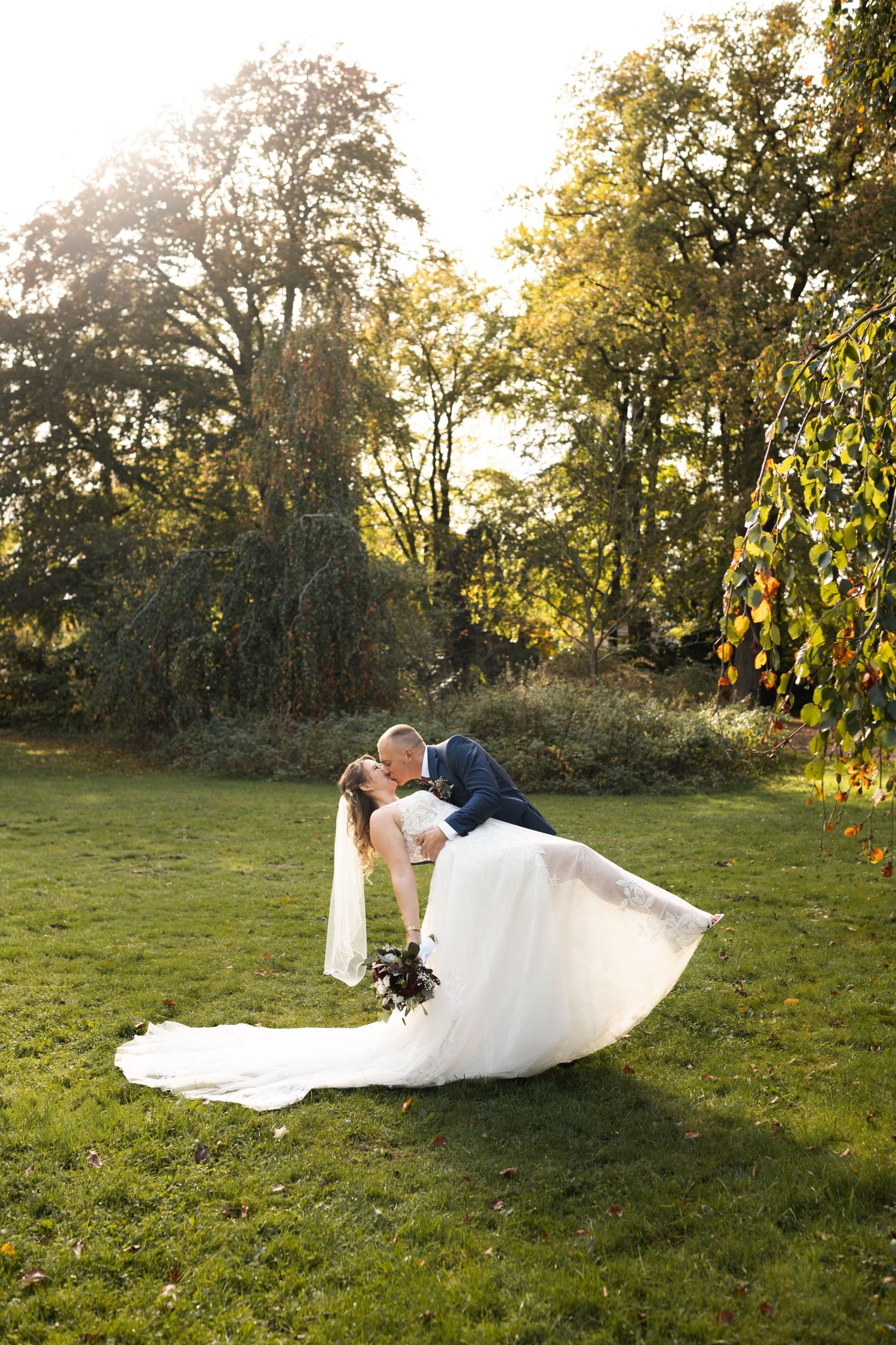 A couple in wedding attire shares a romantic dip pose on a sunlit grassy lawn with autumn trees in the background.
