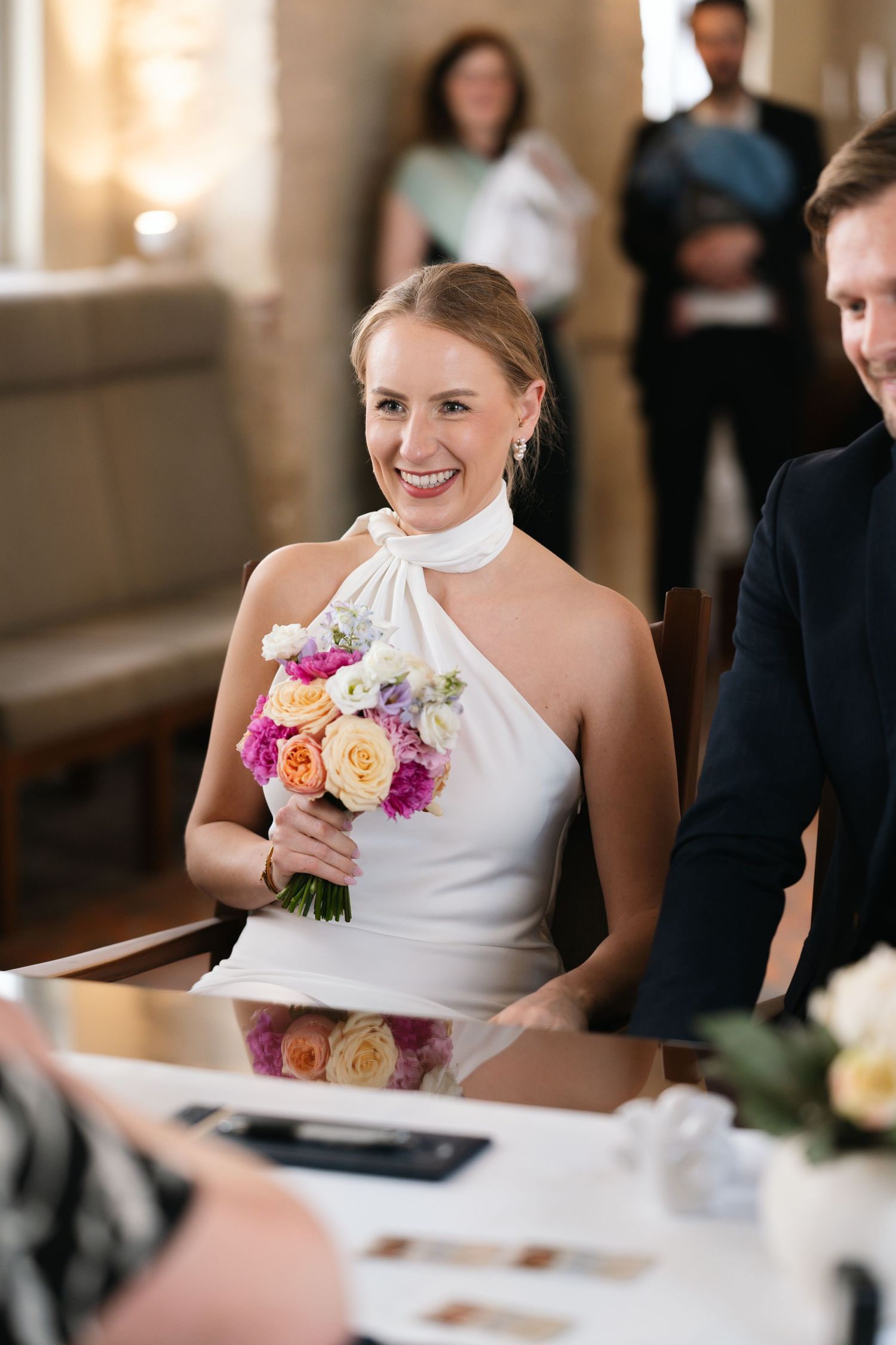 Smiling bride in white halter dress holding colorful bouquet of roses at wedding ceremony table.