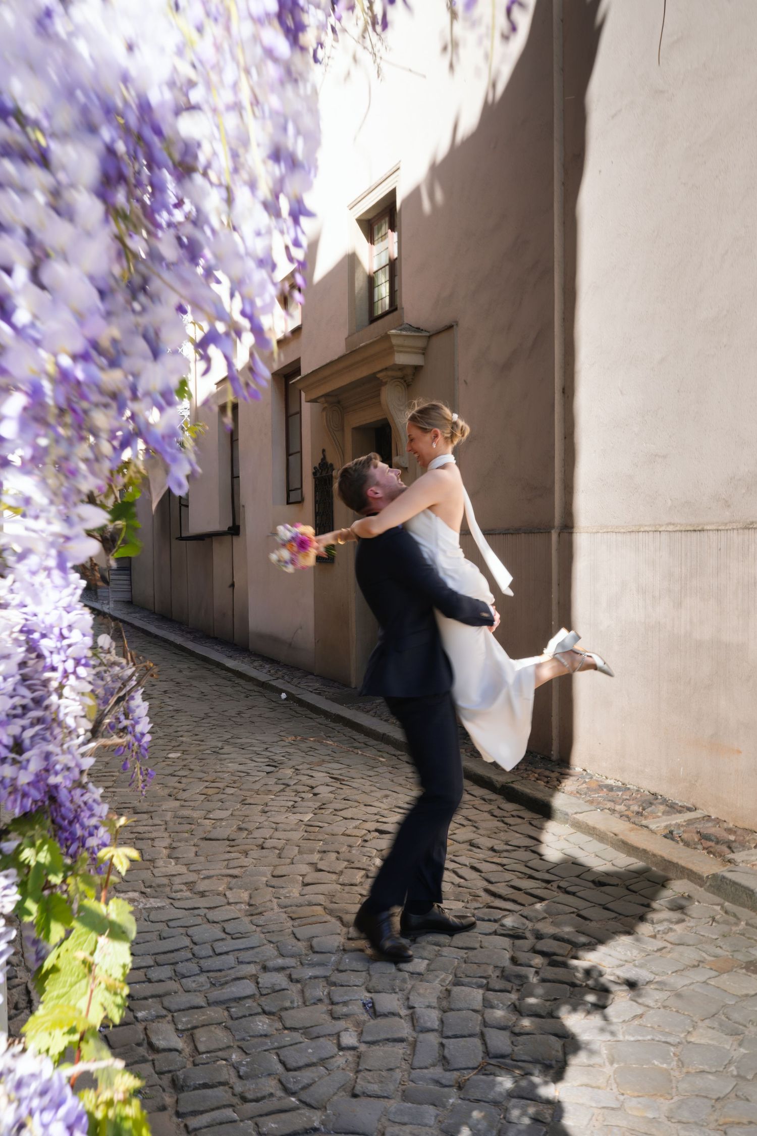 Groom lifting bride in white dress on cobblestone alley with purple wisteria blooms in sunlight.