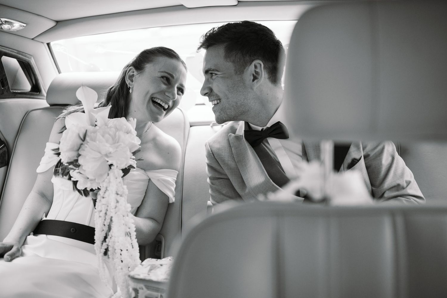 Black and white photo of a smiling bride and groom sharing a joyful moment together in the back of a wedding car.
