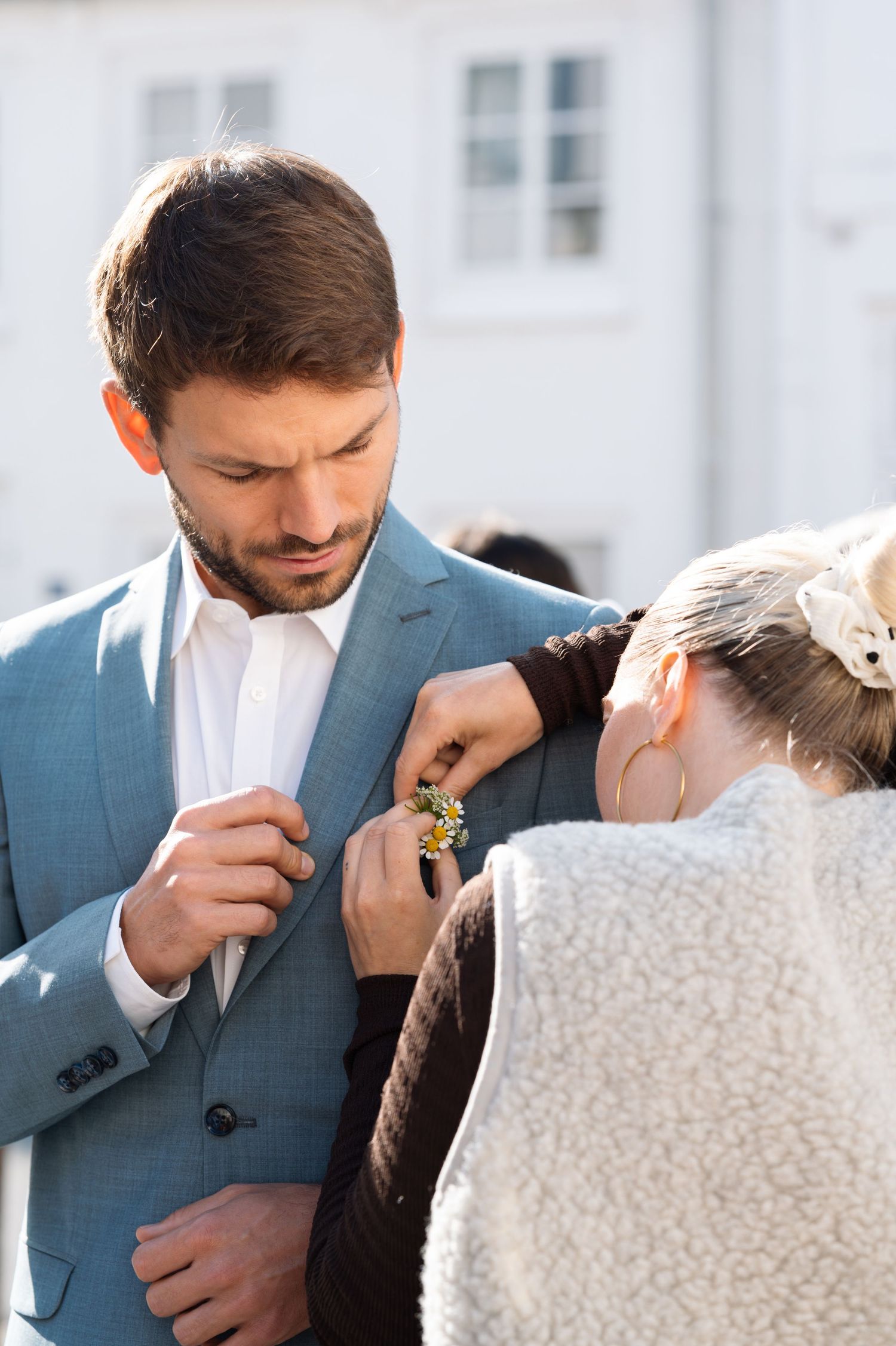 A woman pins a boutonniere on a man in a light blue suit at a wedding ceremony outdoors.