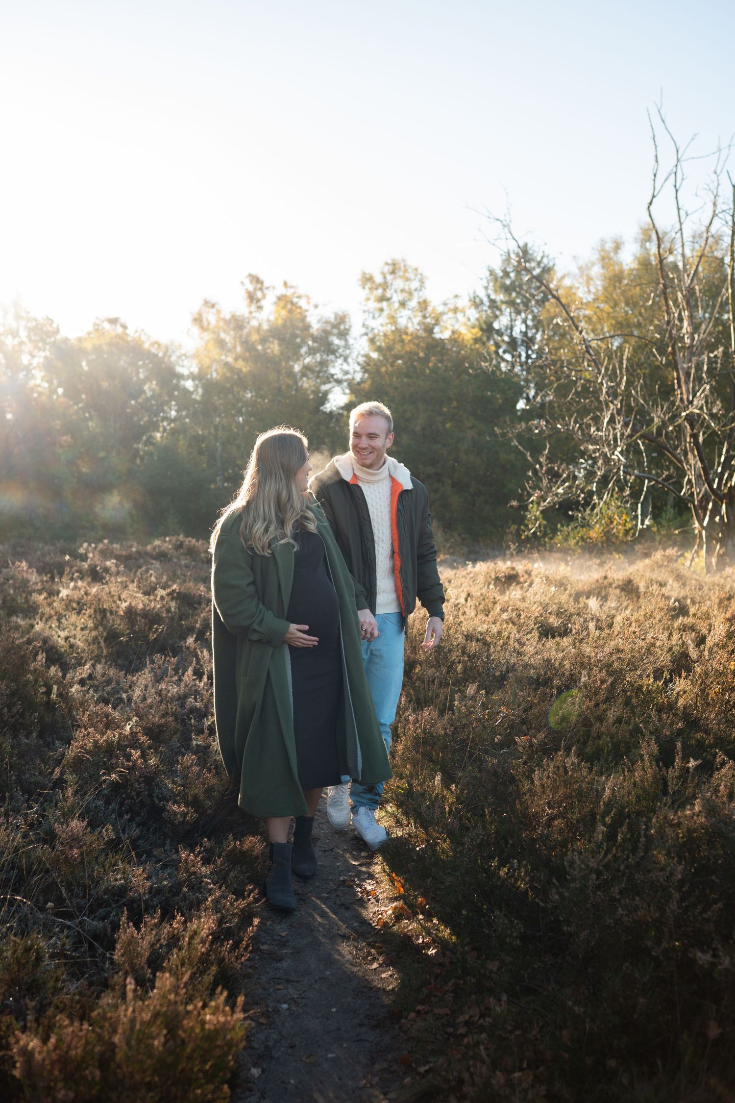 A couple walks along a nature trail during golden hour as sunlight filters through trees and brush.