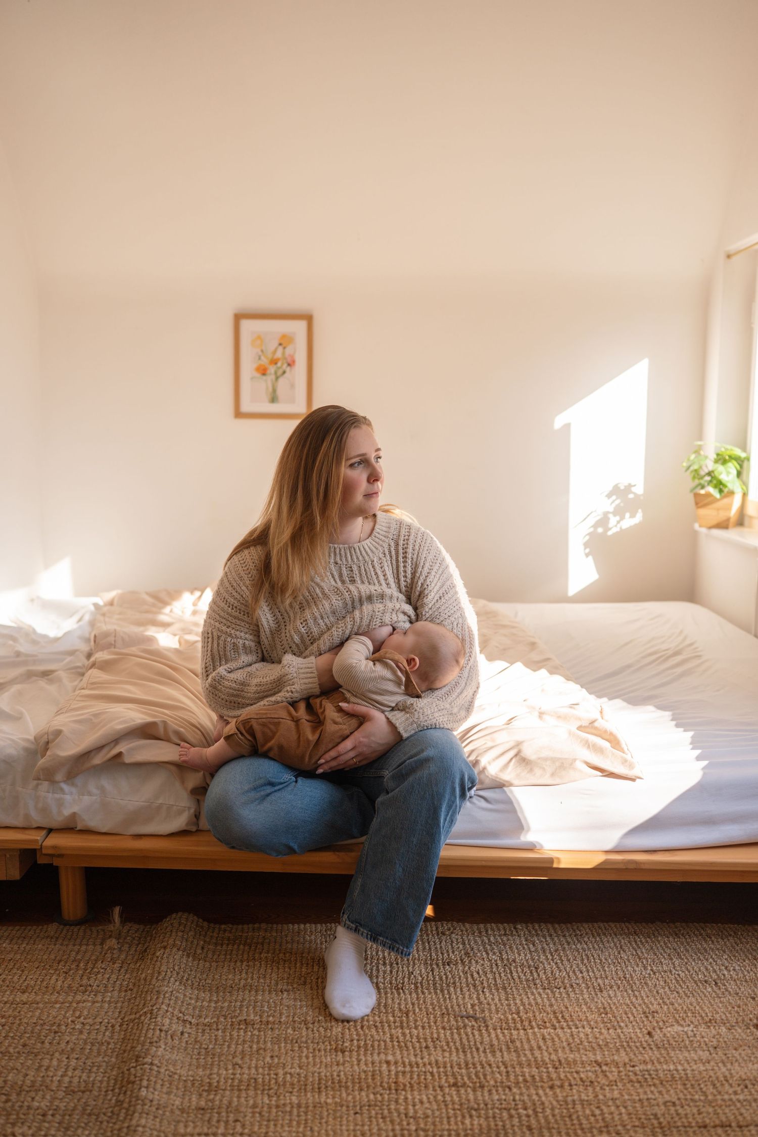A mother sits on a bed holding her newborn baby in warm sunlight, wearing a beige sweater and jeans in a cozy bedroom.