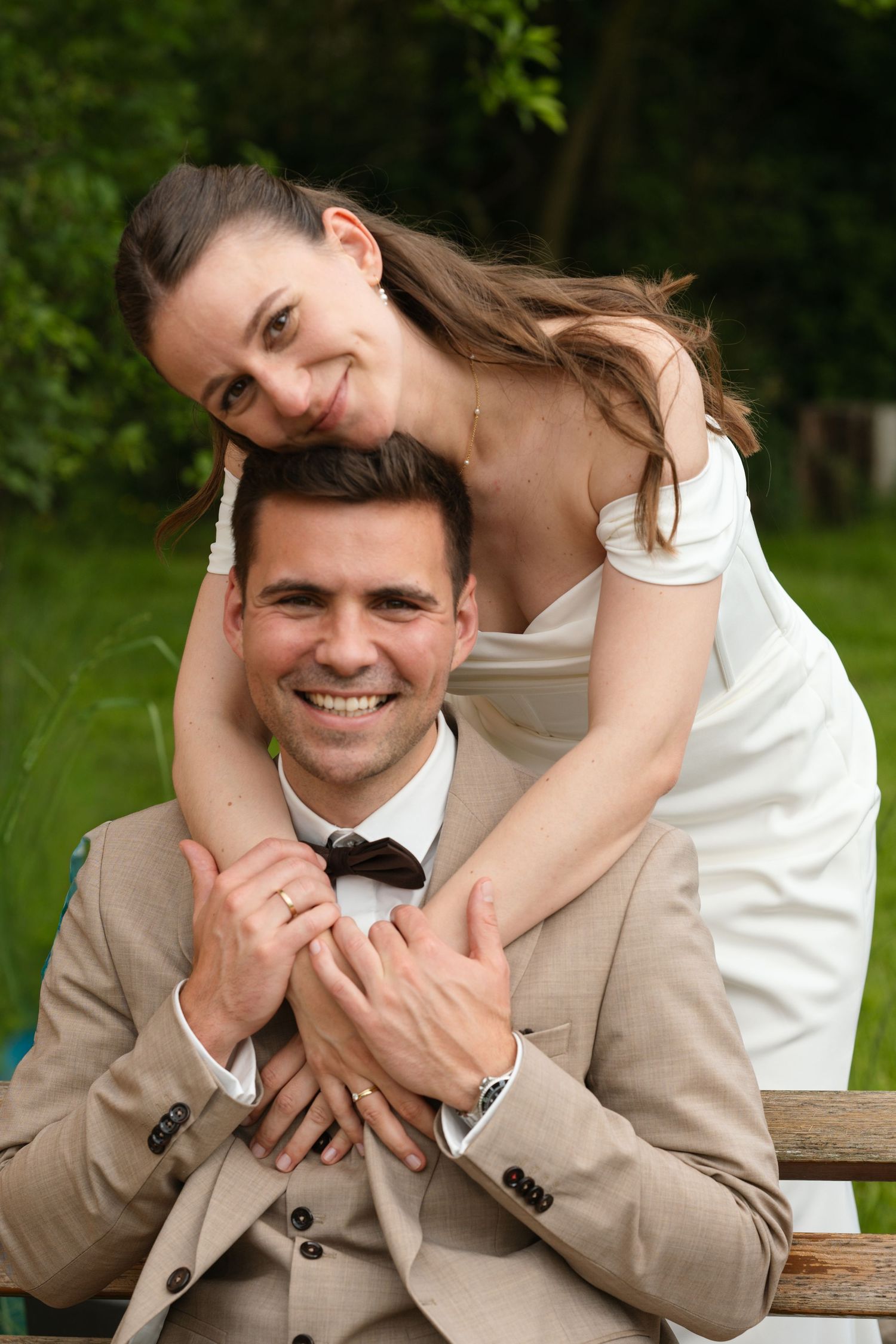 Smiling bride in white dress embraces groom in tan suit with bow tie on a bench outdoors with green trees.