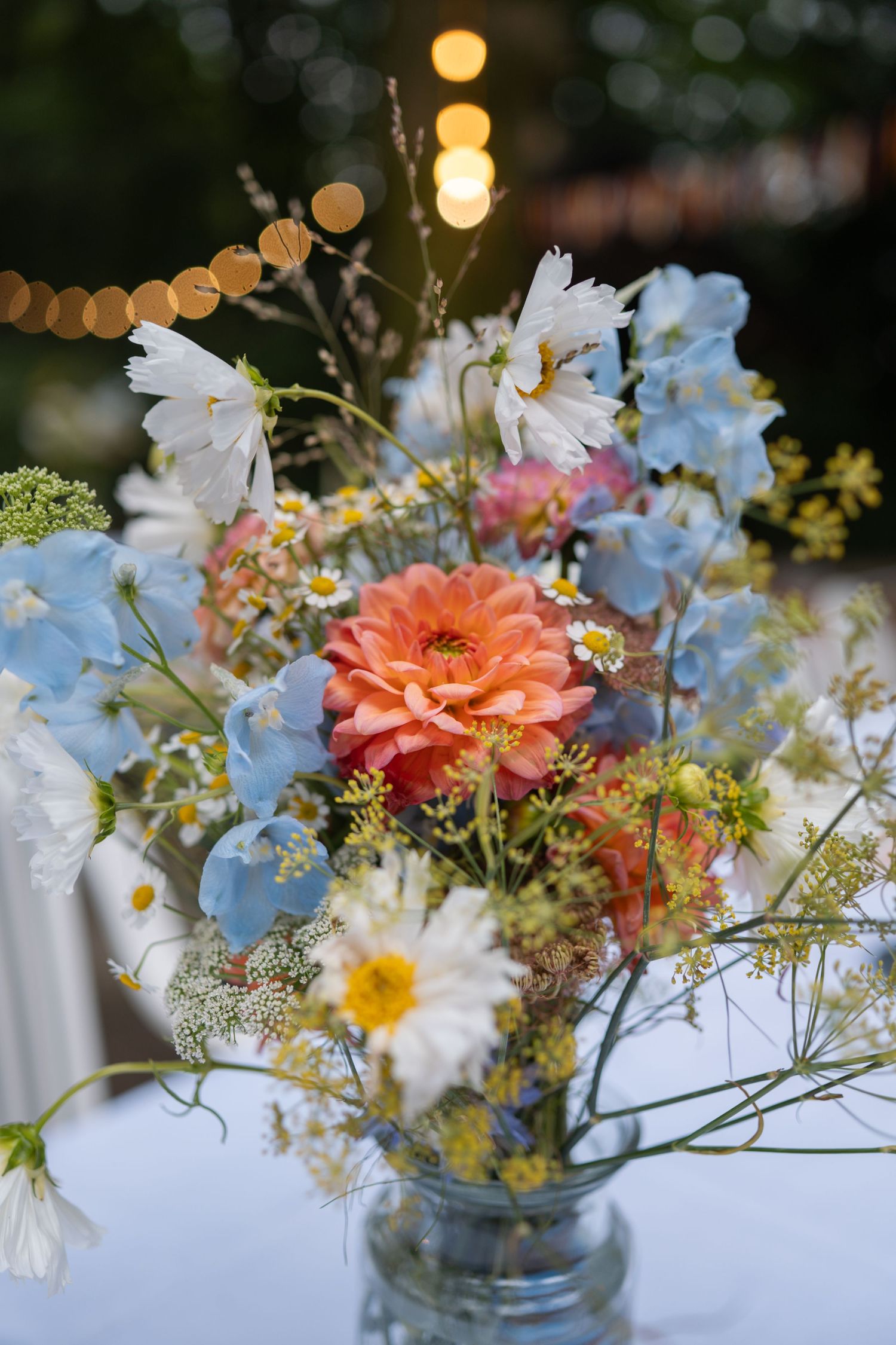 A vibrant floral arrangement in a glass vase featuring peach dahlias, blue delphiniums, white cosmos, and daisies on a table.