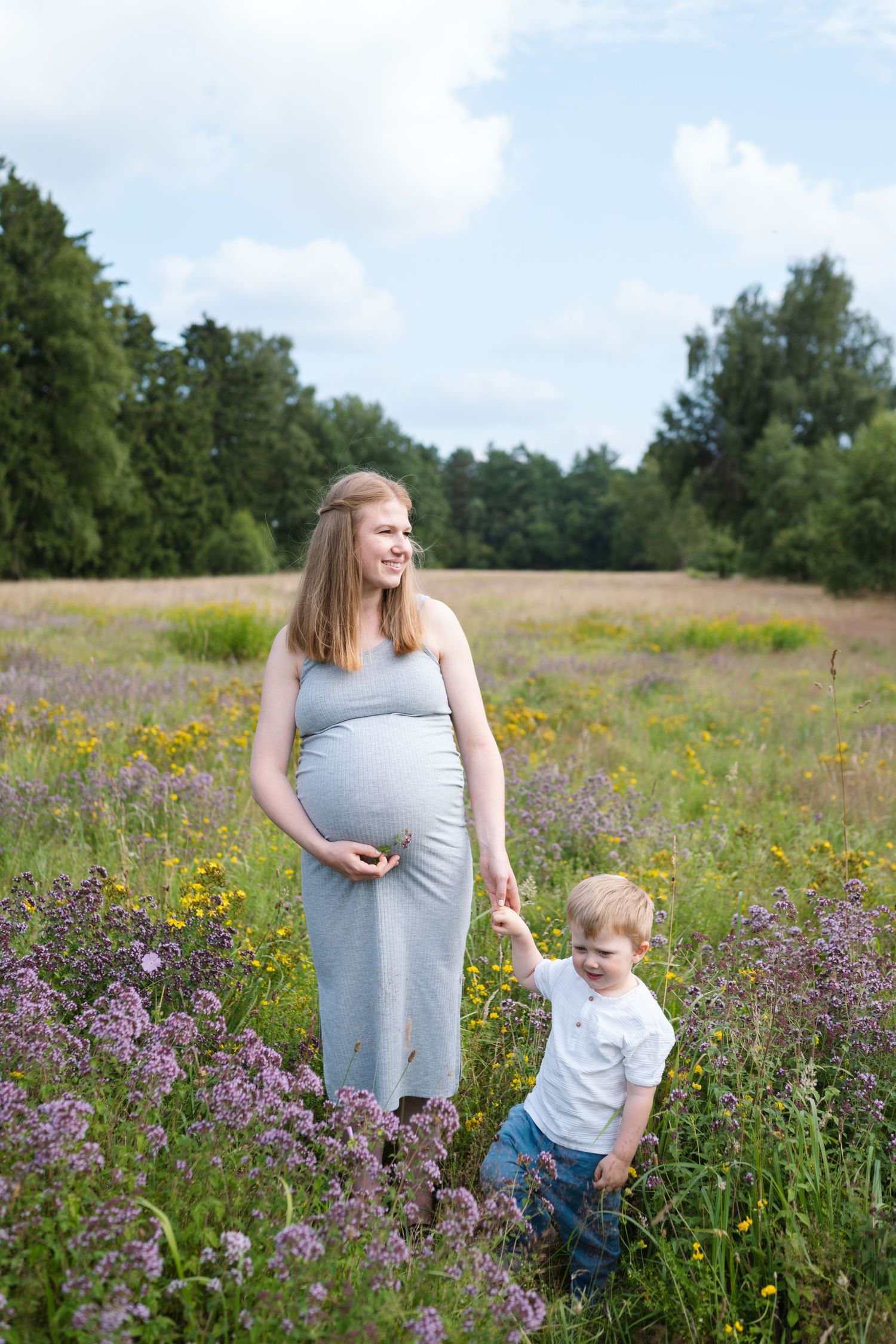 A pregnant woman and toddler explore a vibrant meadow filled with purple wildflowers and yellow blooms on a sunny day.