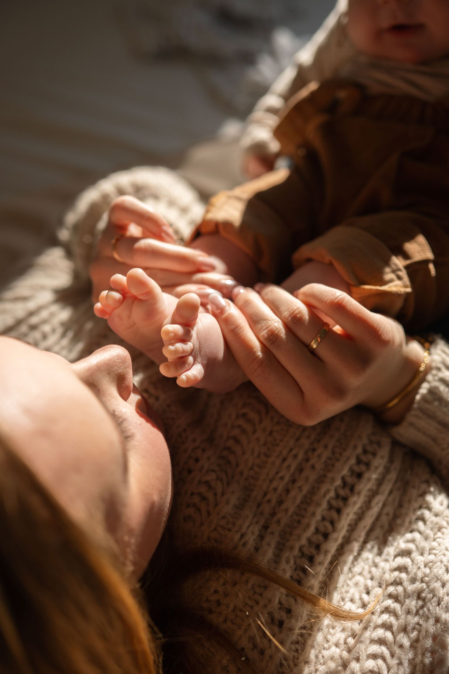 A mother lying down holds her newborn baby's tiny feet and hands, wearing a knit sweater and gold ring.
