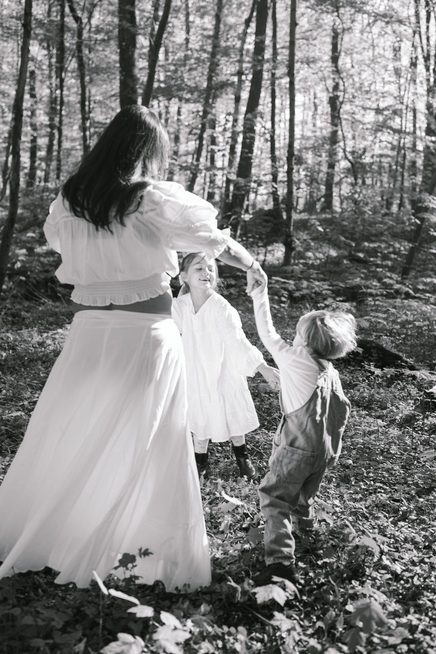 Black and white photo of a mother in white dress dancing with two young children in a sunlit forest.