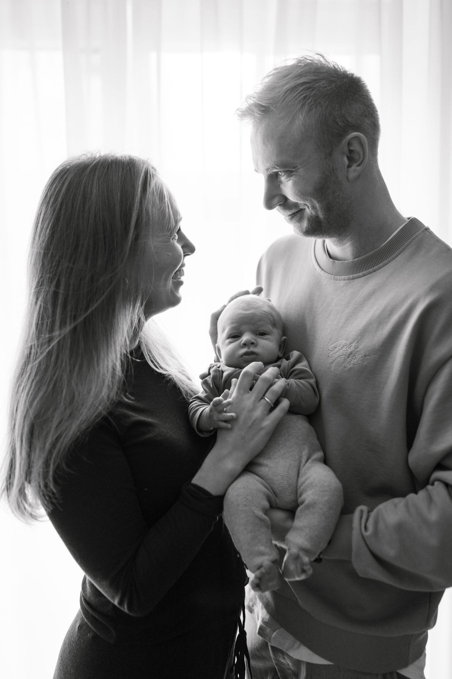 Black and white photo of smiling parents holding their newborn baby near a bright window.