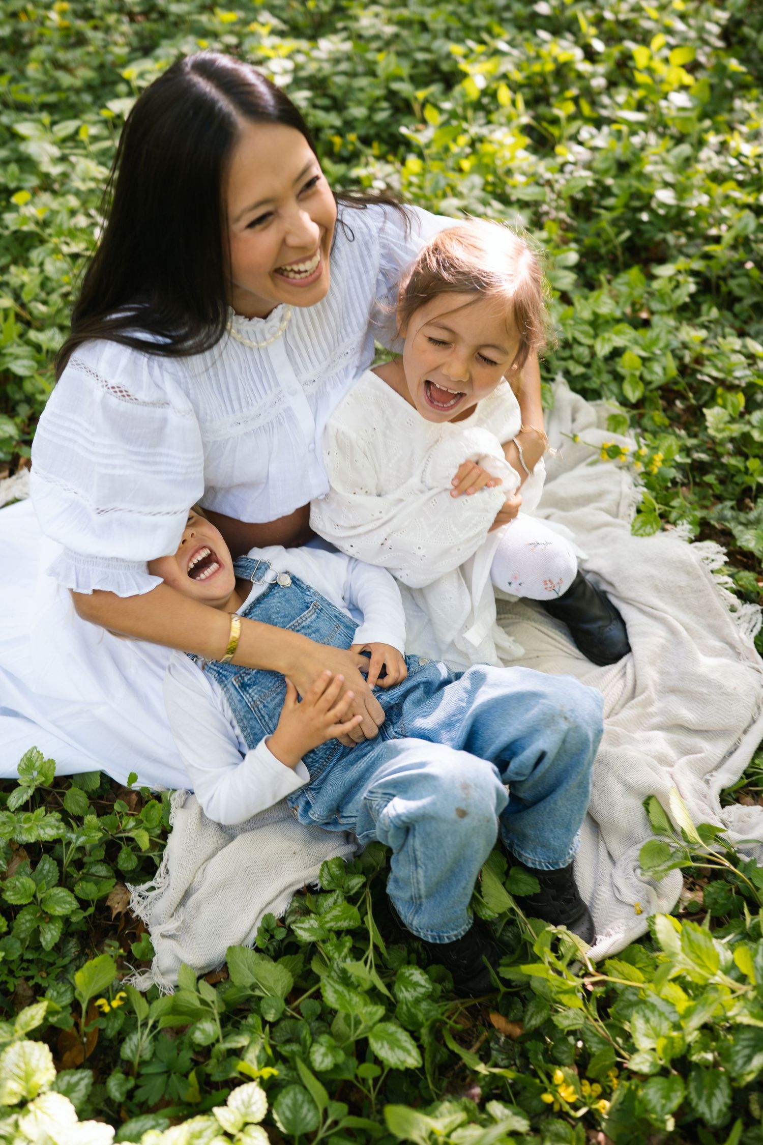 A joyful mother in white dress laughs with two children in blue jeans while sitting in lush green garden.