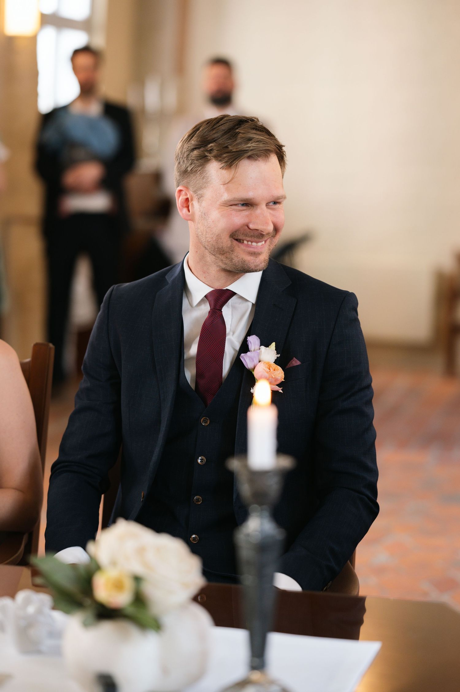 Smiling groom in navy suit with floral boutonniere seated at wedding ceremony table with candles.