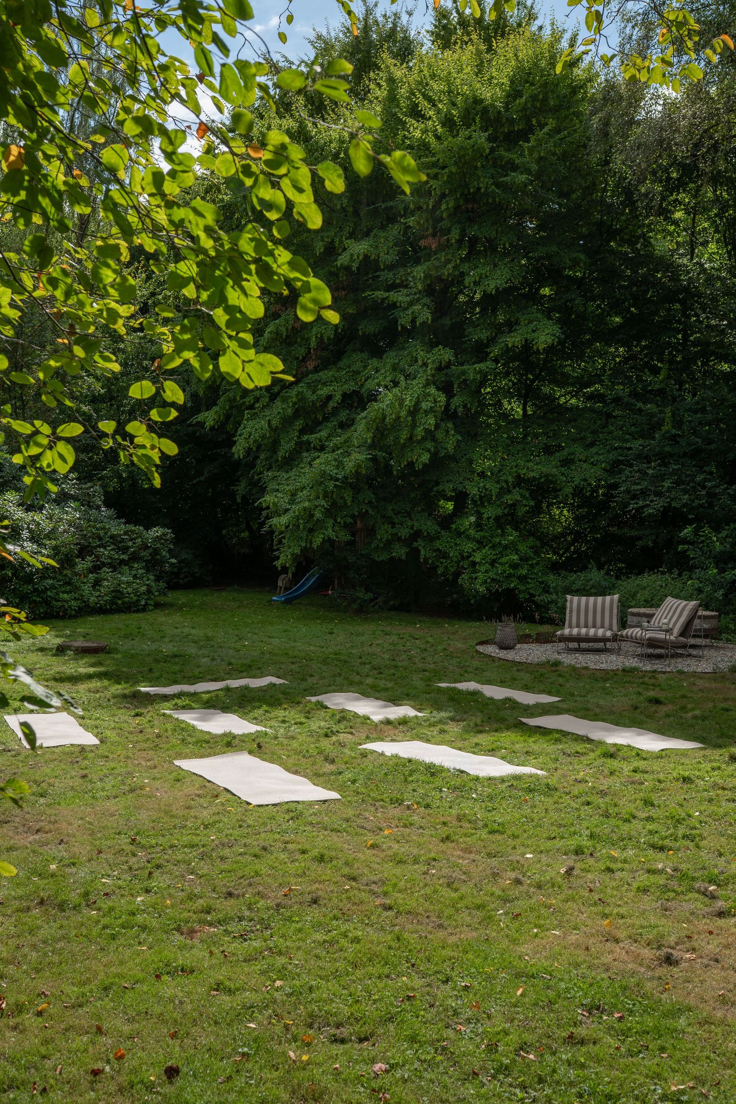 Peaceful garden setting with yoga mats laid out on grass surrounded by lush green trees and wooden benches.