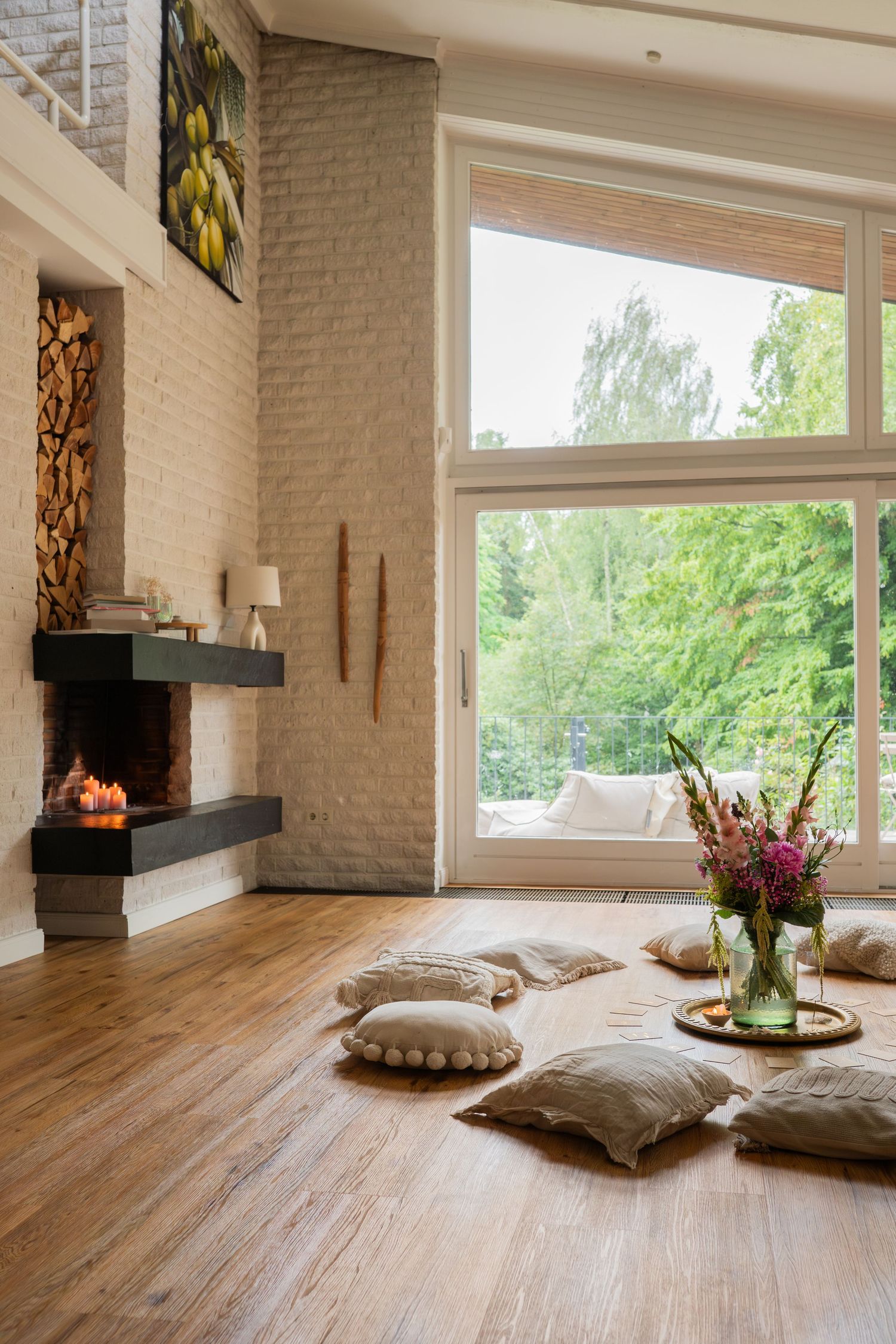 Modern living room with floor cushions, fireplace, large window view of trees, and fresh flowers in a vase on wooden floors.