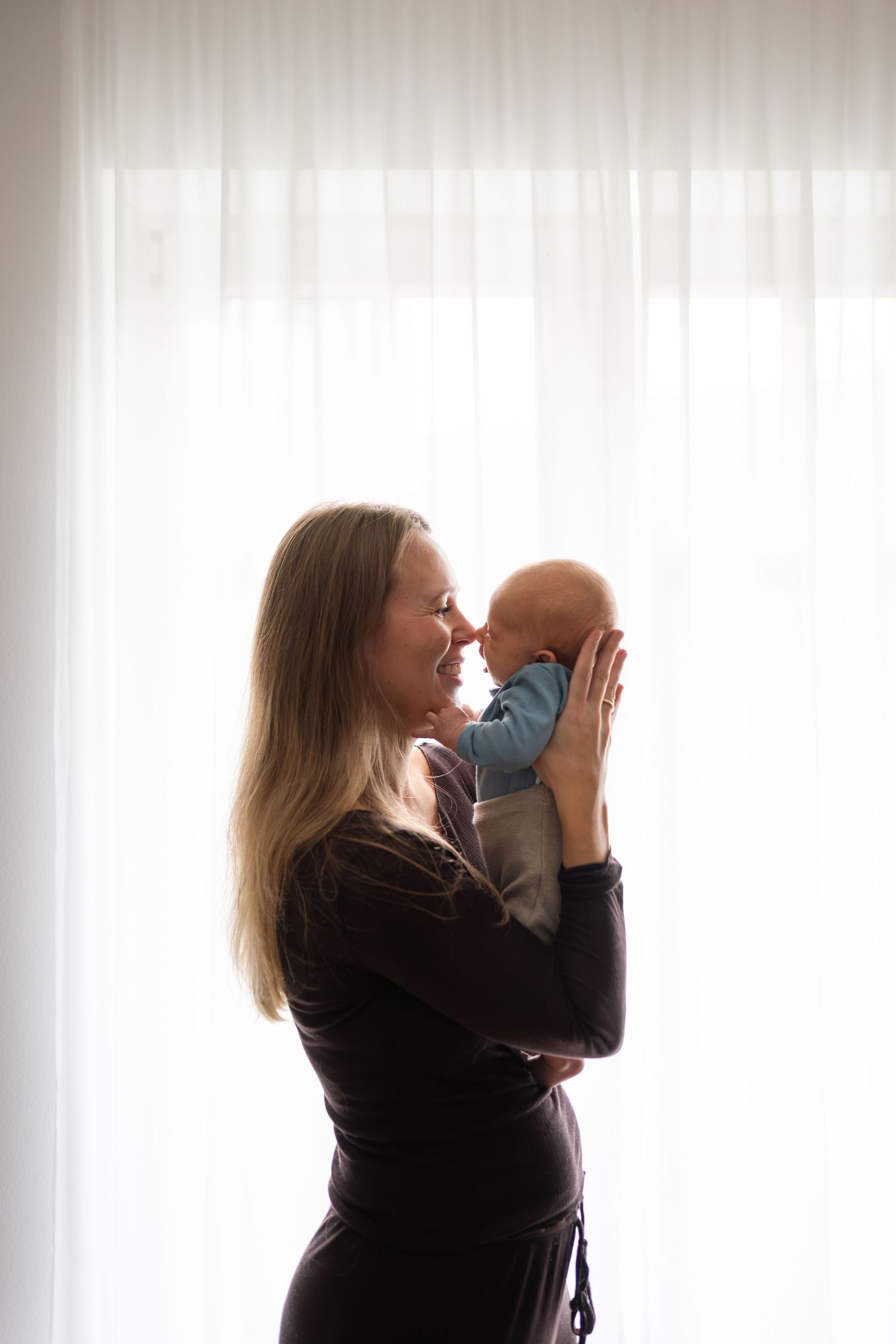 A smiling mother holds her newborn baby up to her face near a bright window, sharing a tender moment.