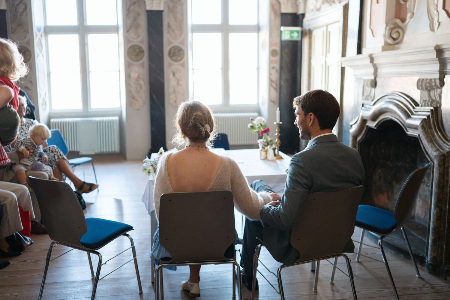 Couple seated at a civil wedding ceremony in an elegant room with guests watching from chairs nearby.
