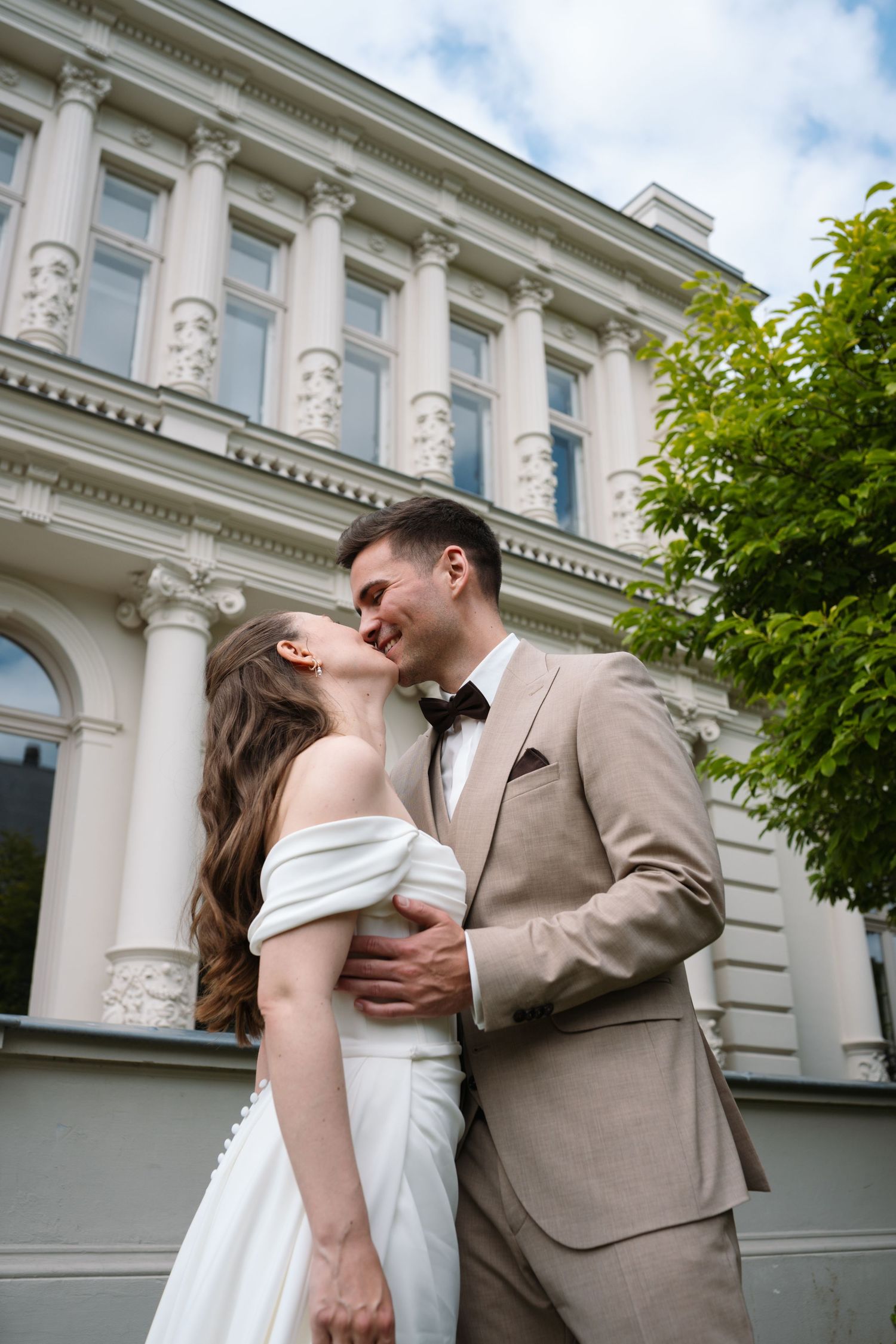 Bride and groom share a romantic kiss outside an elegant white classical building on their wedding day.