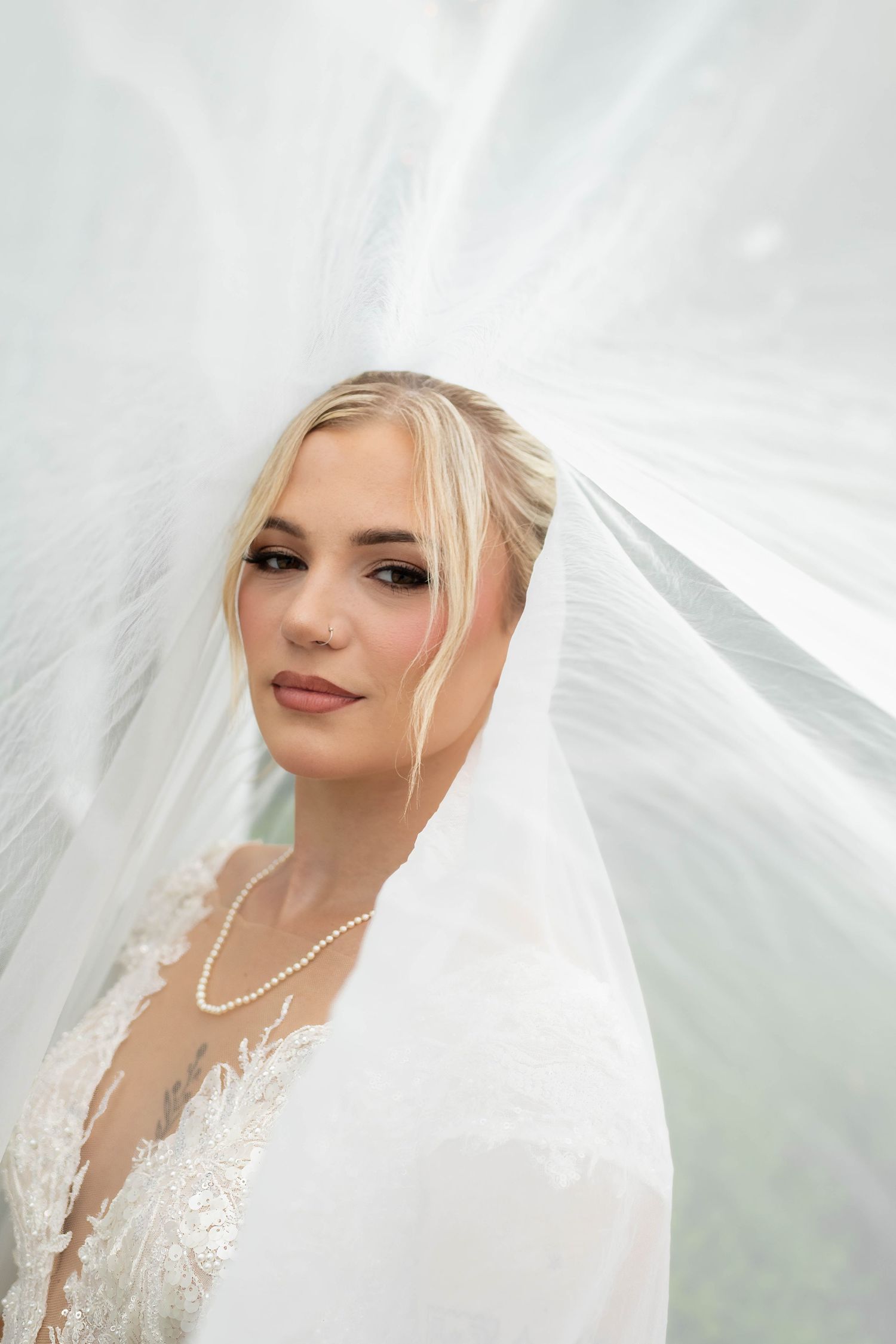 Bride with blonde hair and pearl necklace draped under flowing white veil on wedding day.