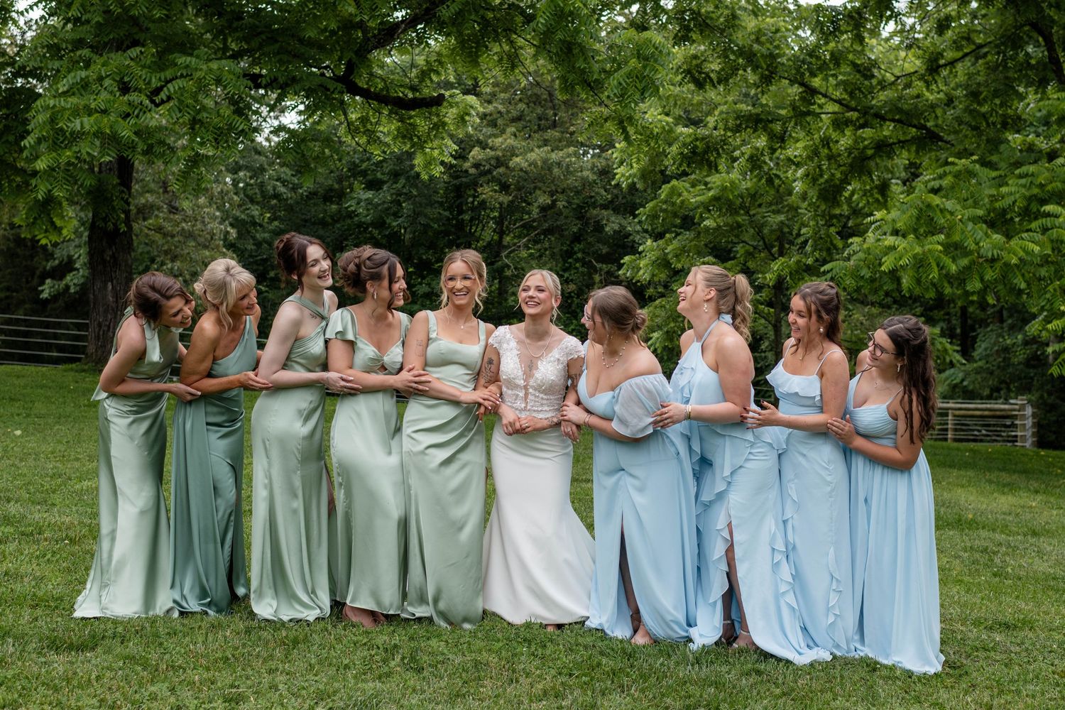 Bride in white lace gown laughing with bridesmaids in sage and blue satin dresses outdoors.