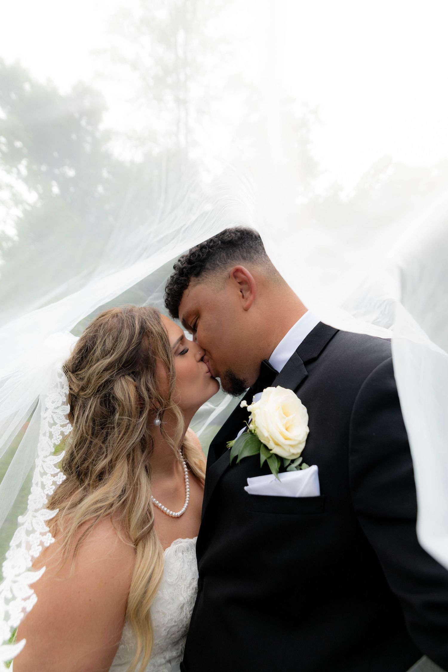 Bride and groom sharing a romantic kiss under flowing white veil in soft natural light.