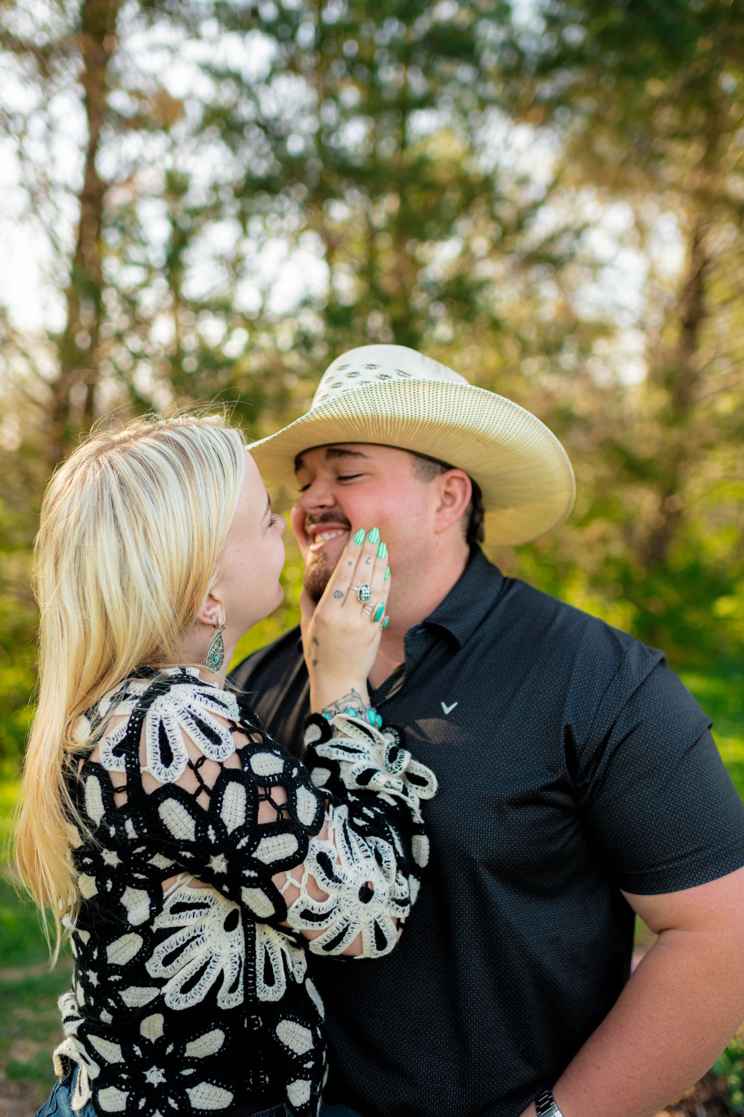 Woman playfully touching smiling mans face outdoors, man wearing a straw cowboy hat in green setting.